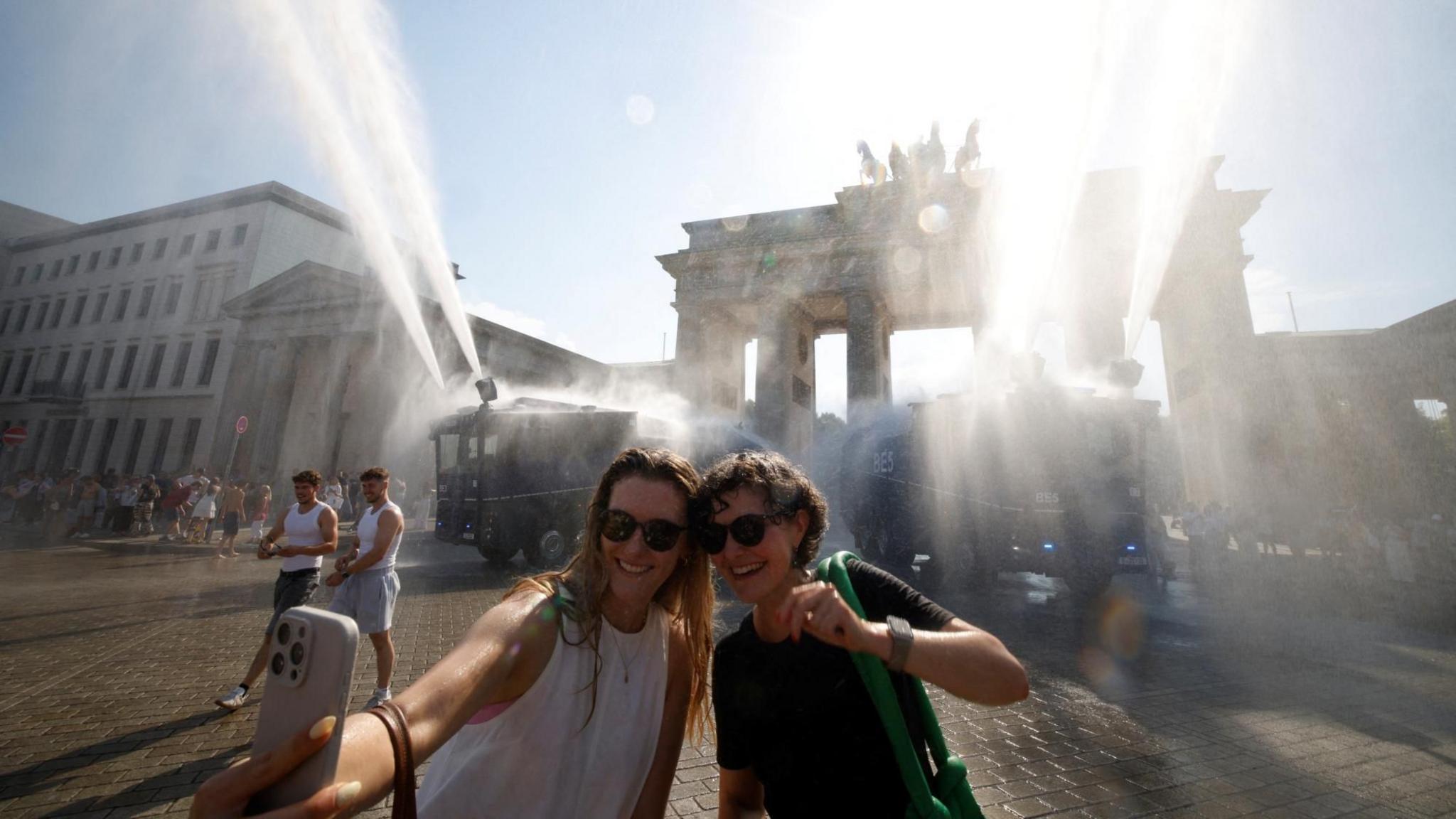 Two women take a selfie as they stand under mist fired from two police water cannons