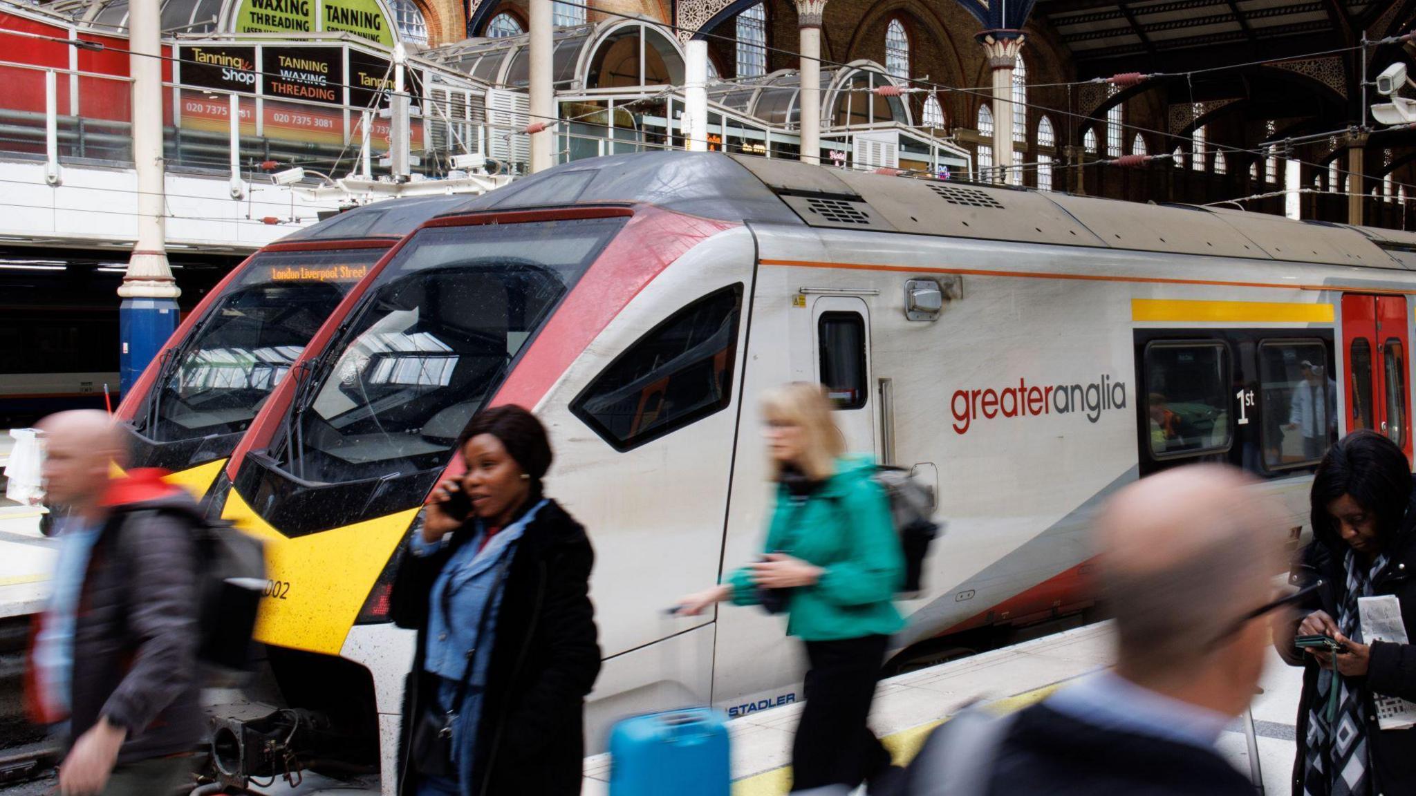 People at Liverpool Street station with a train on a platform. Some are on mobile phones, others have luggage.