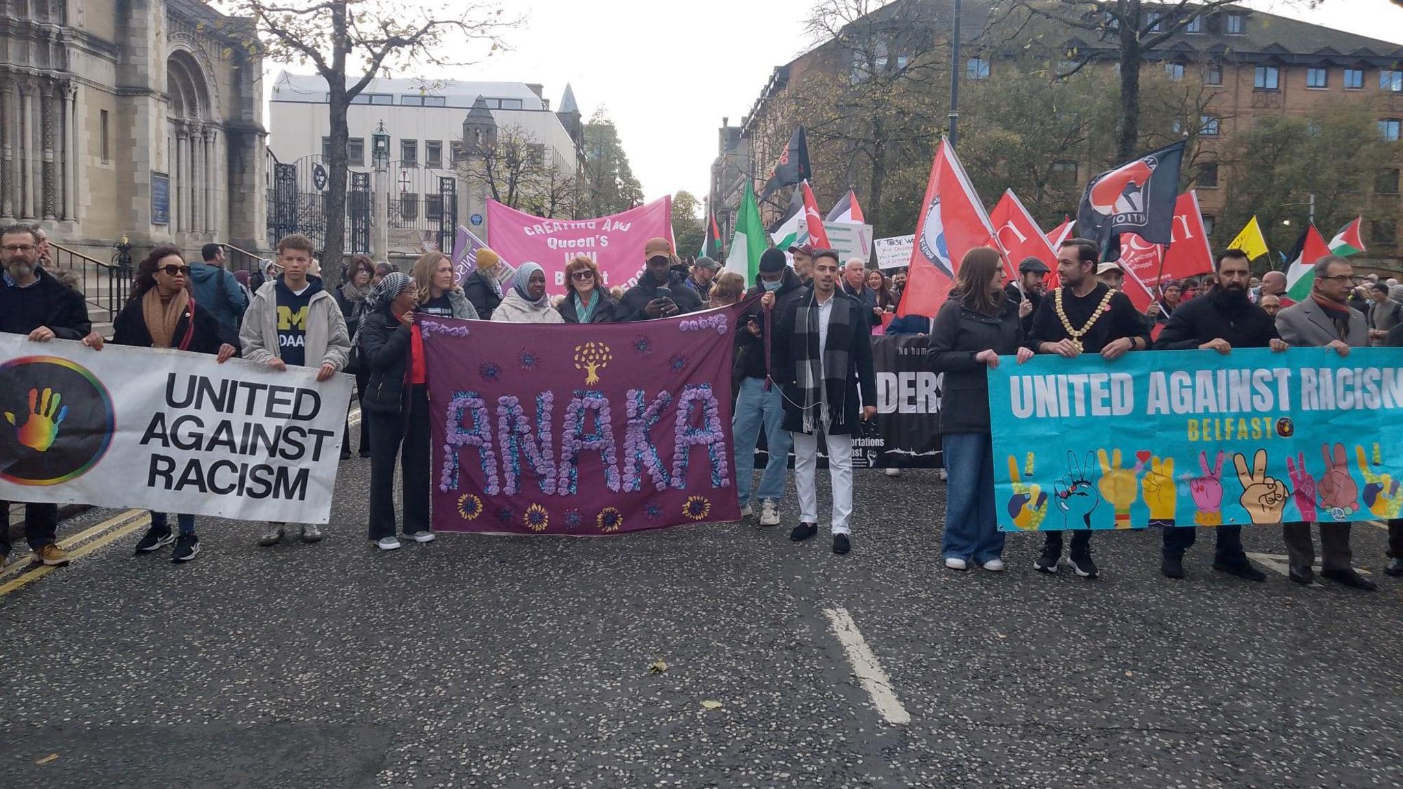 A wide shot of protestors holding banners, including United Against Racism and Anaka as they walk down the street. Many are holding flags, including trade union flags and the Palestinian flag.