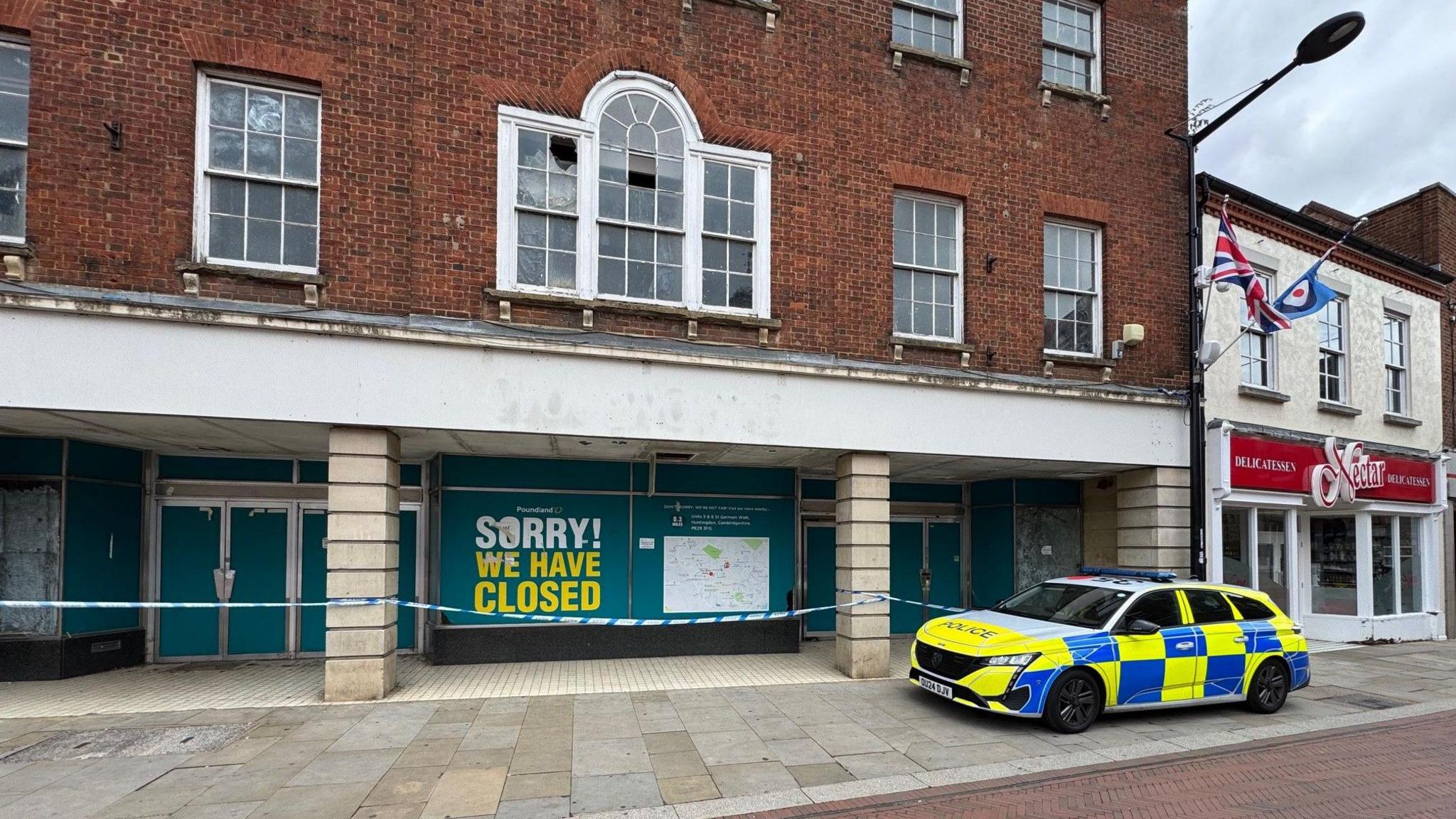 A police car outside the old Poundland store. There is police tape on the front of the building and a large sign on the shop that says 'sorry we have closed'. A window has been smashed on the upper floor.