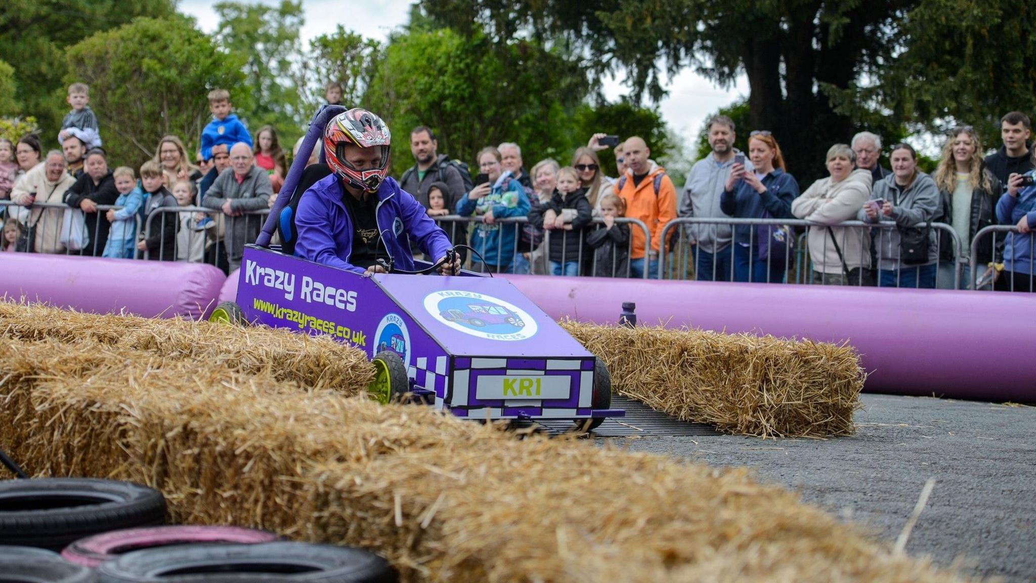 A man in a small purple go-kart that is racing down a road. The kart has white writing on it that reads "Krazy Races". Tyres and hay bales are also on the road. A crowd is gathered to watch the race behind metal barriers.