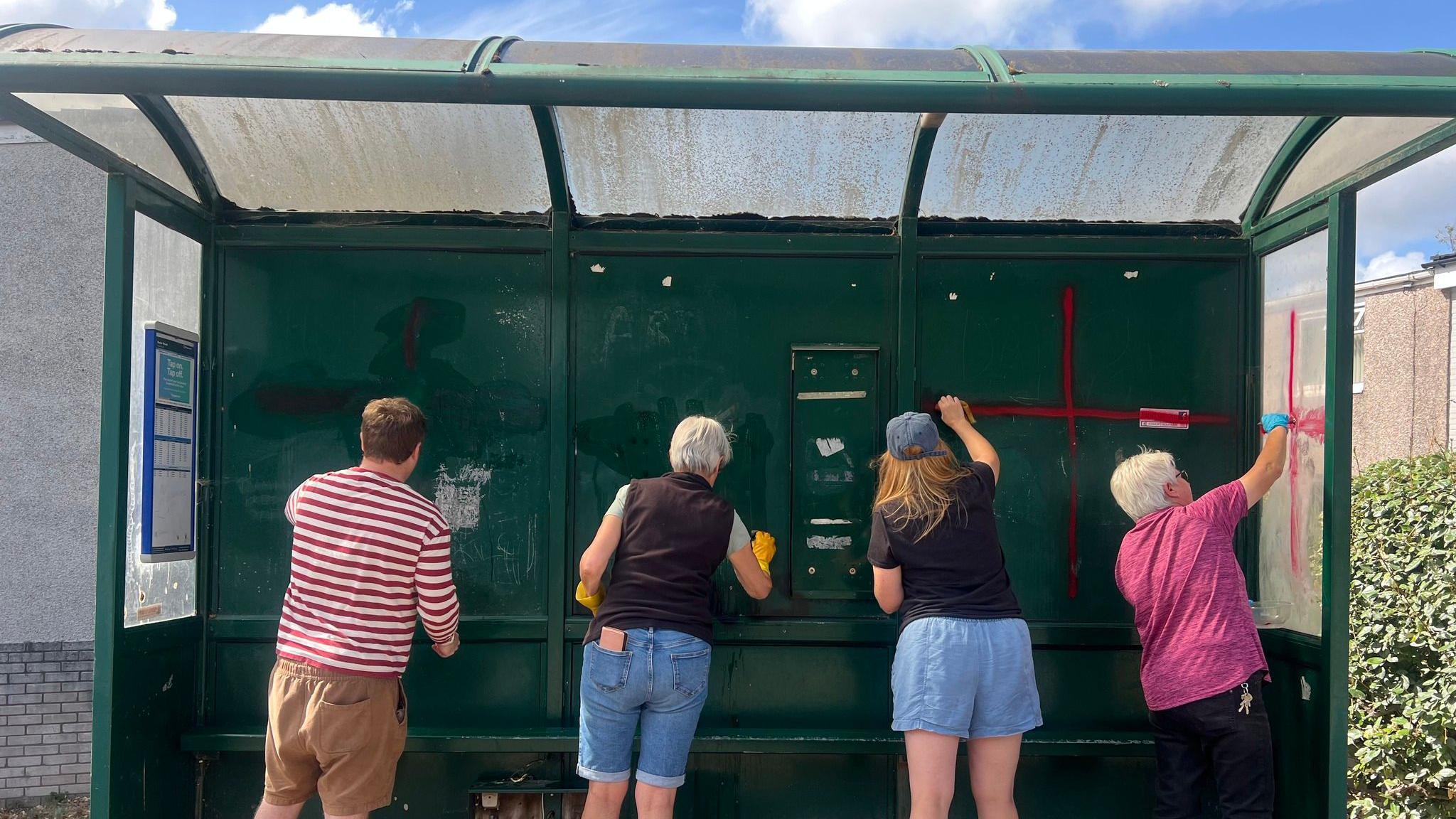 Four residents photographed with their backs to the camera inside a dark green bus stop scrubbing off paintwork. Some wear yellow rubber gloves. A red cross can be seen on the back of the bus station, as well as one on the right hand side plastic window pane.