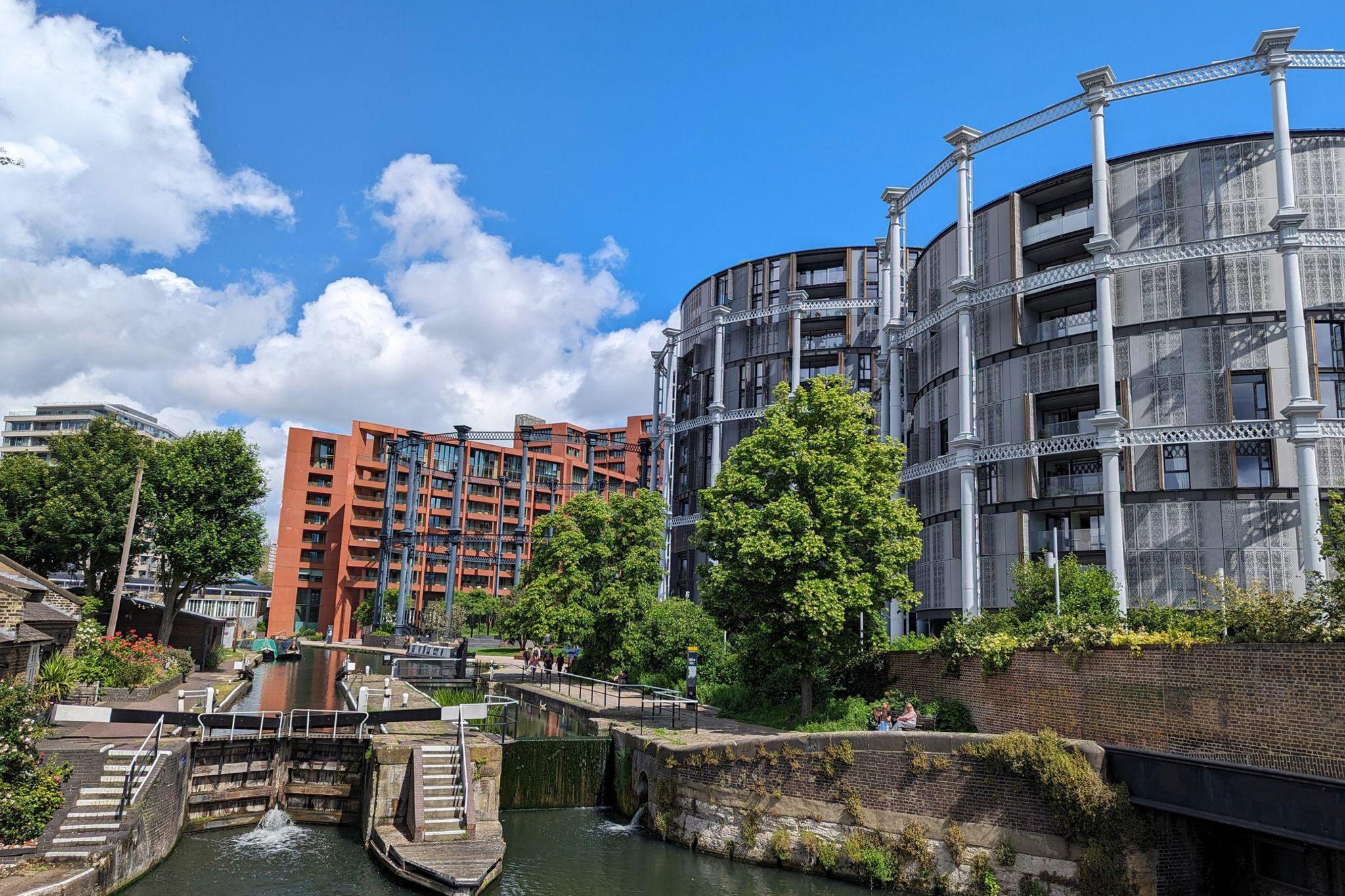 New flats built alongside a canal lock in London. Two round apartment blocks are built inside old gas holders, whilst a red-brick block of flats is visible in the background. There are lots of trees and a towpath lining the canal.