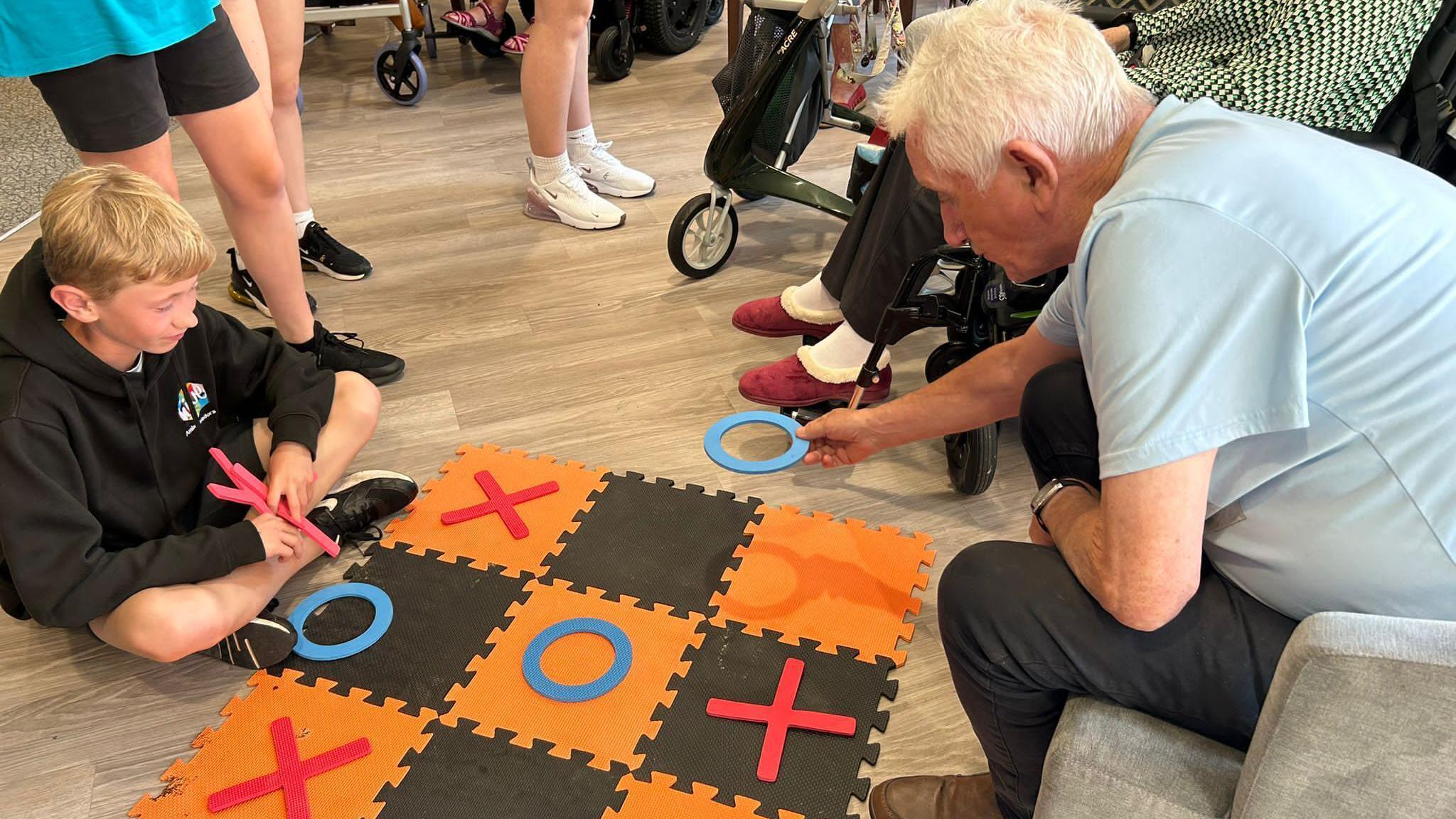 A teenage boy with blond hair and wearing a black hoodie plays noughts and crosses on a large board on the floor with a man who has grey hair and is wearing a light blue t-shirt.