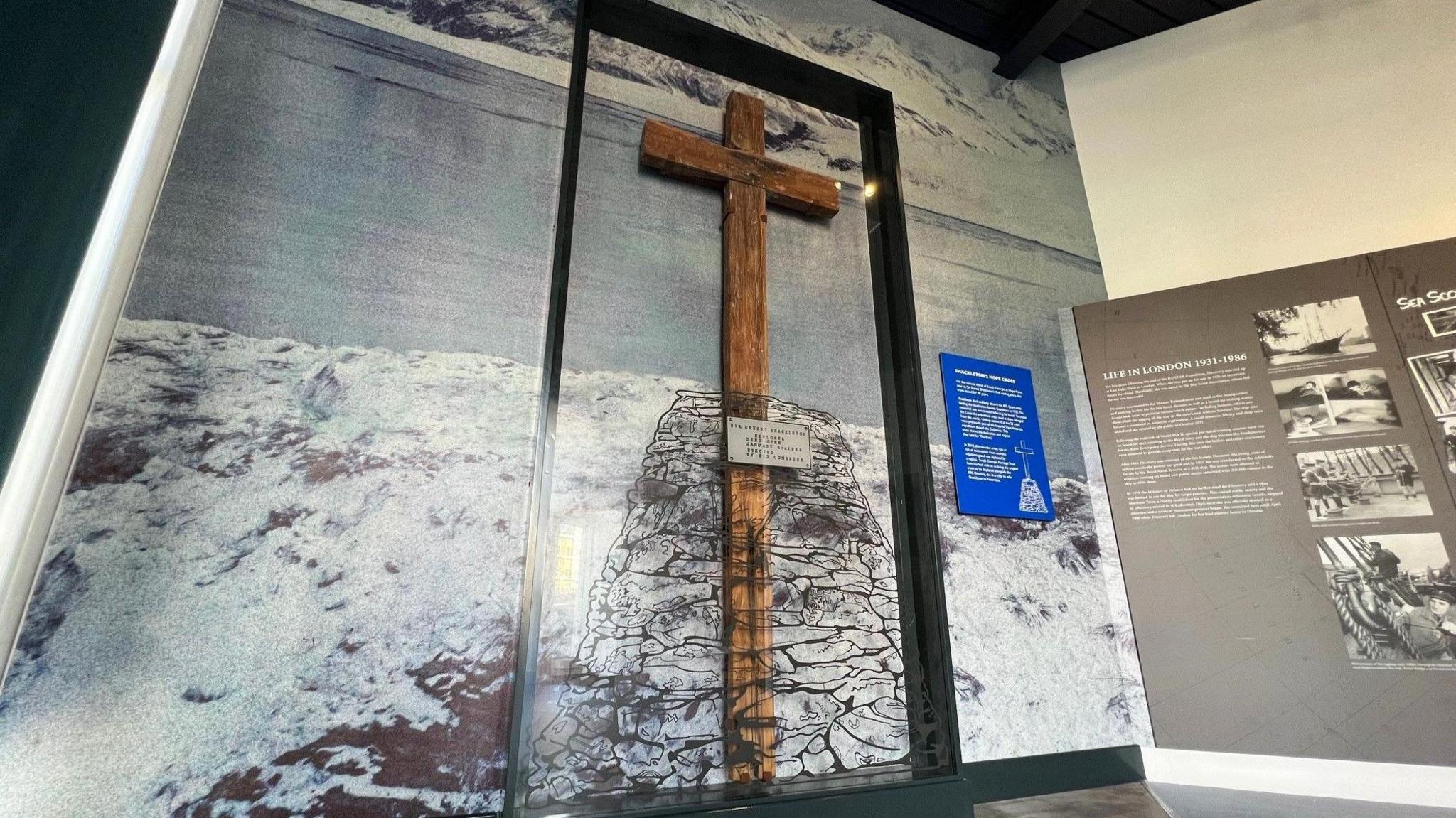 A large wooden cross behind perspex in a black frame mounted on a wall in front of a picture of the Antarctic