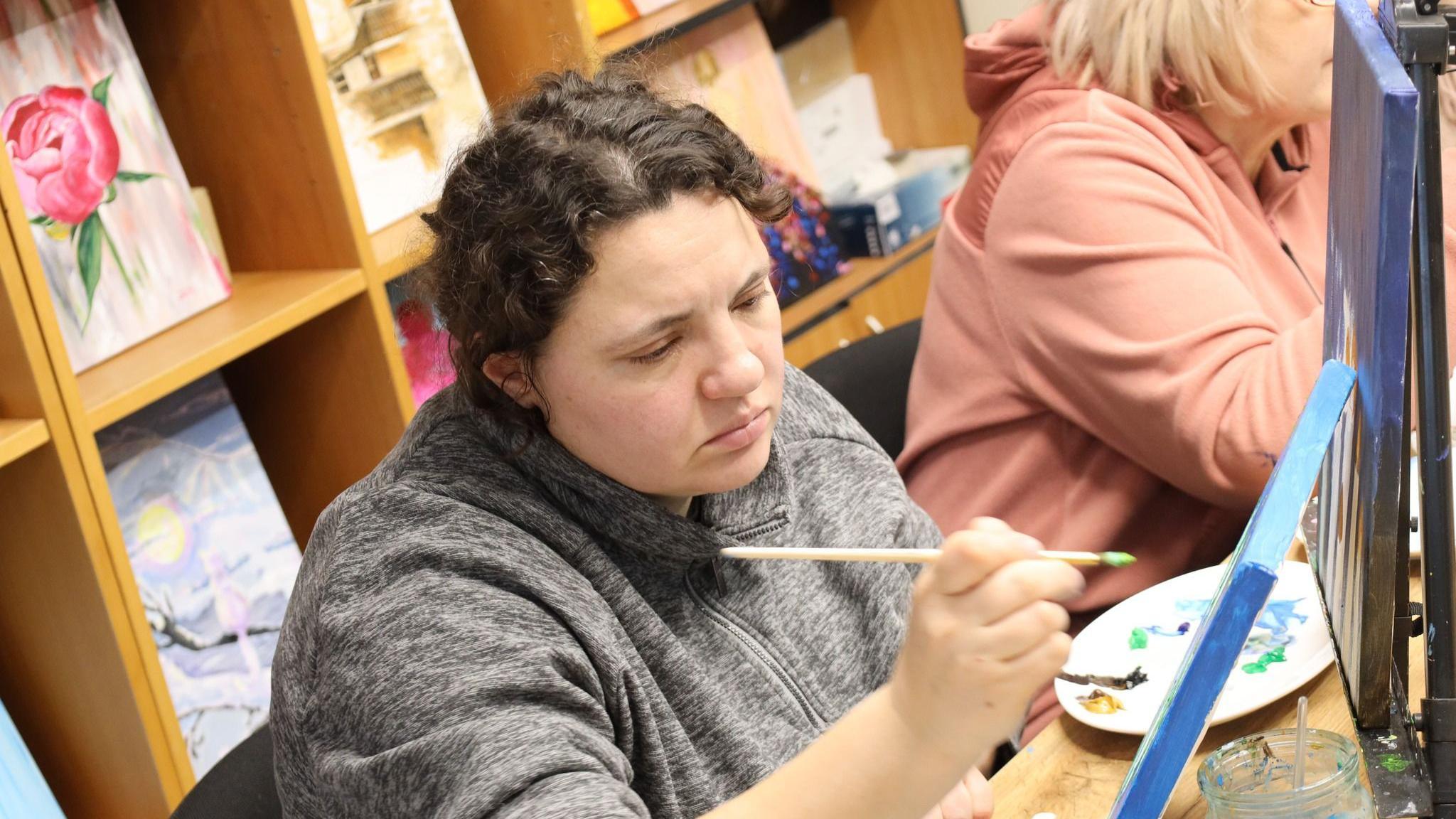 A woman with curly brown hair wearing a grey fleece paints.