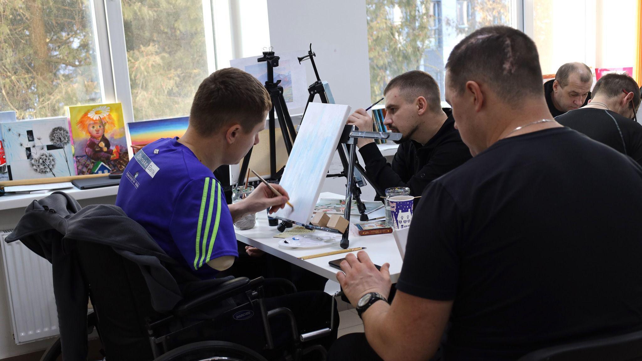 A man with his right arm missing sits in a wheelchair painting. He is wearing a blue and green football shirt. 