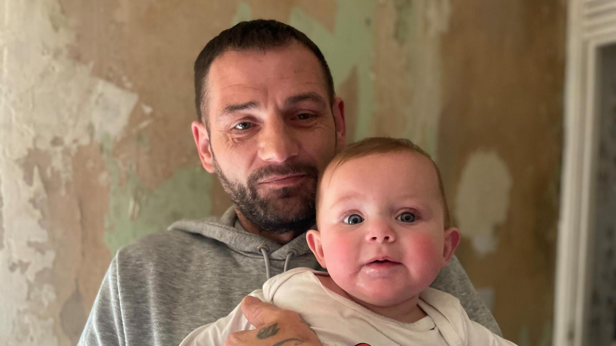 A man with closely cropped hair and a beard holds a baby up to the camera. Behind them the wall is bare.