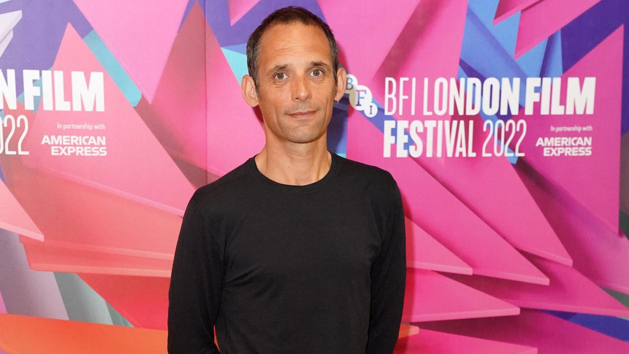 Mark Jenkin is smiling at the camera wearing a long-sleeved black top. He has short dark hair and brown eyes. He is standing in from of a pink and blue display which reads: BFI LONDON FILM FESTIVAL2022 sponsored by AMERICAN EXPRESS.