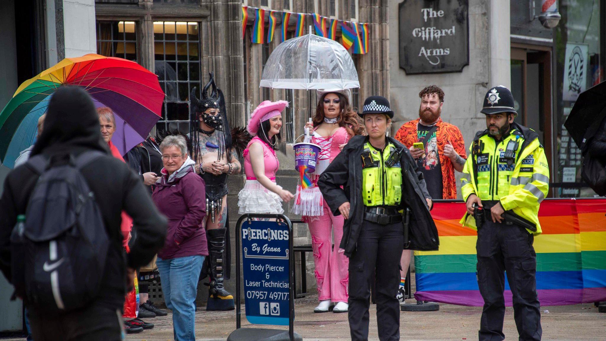 Two police officers dressed in uniform and wearing helmets, stand watching on. Behind them is a rainbow-coloured barrier. People, some dressed  in bright costumes, are standing behind them.