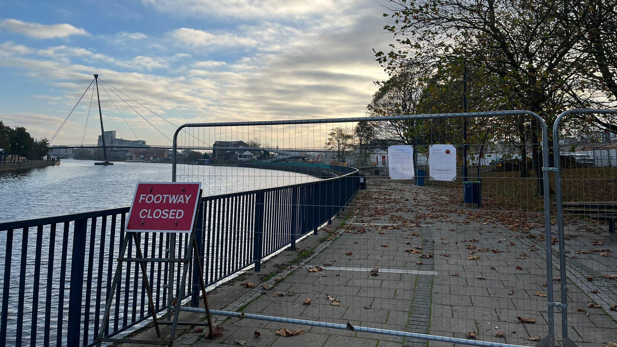 A footpath along the River Tees is blocked off by metal grating. A red sign reading "FOOTWAY CLOSED" is placed in front. A bridge of the river can be seen in the distance.