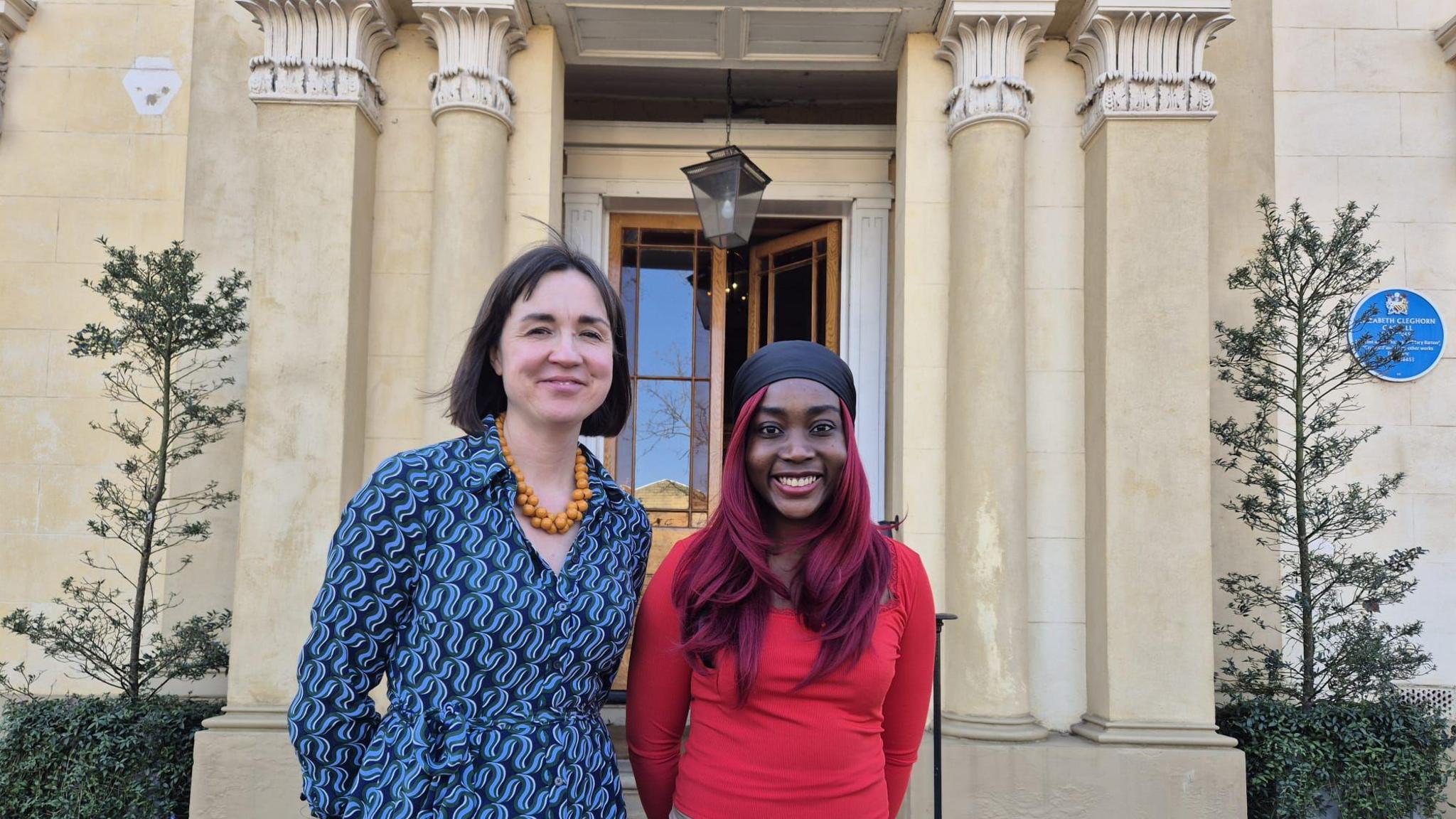 Sally and Princess stand outside the entrance to the cream-coloured stone mansion, which has columns and tall plants on either side and a blue plaque marking the author's achievements on the right