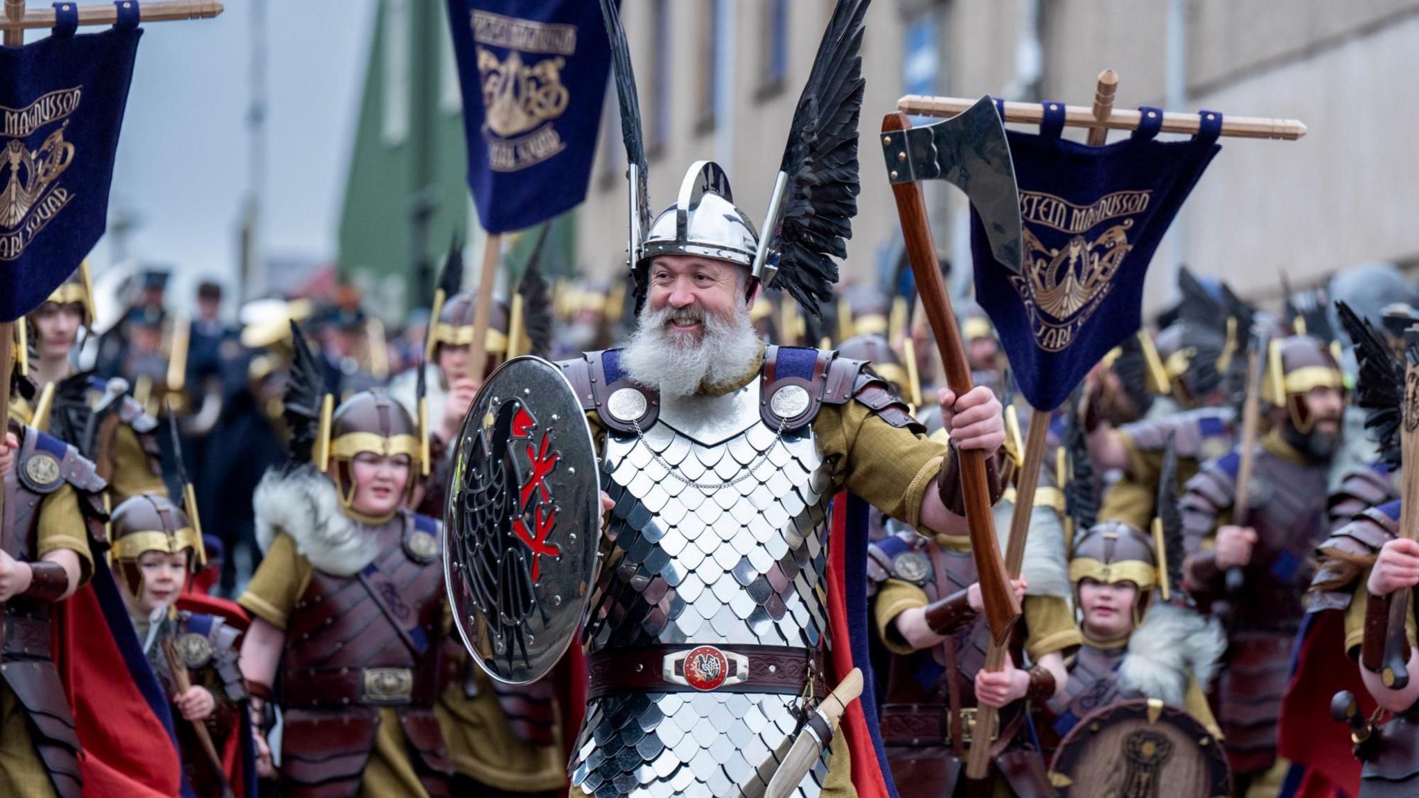 The guizer jarl in full Viking dress. He is holding a shield with a bird insignia and a helmet with metallic wings on the side. He holds aloft an axe. He has a very full grey beard. Behind are more men dressed in Viking costume.  