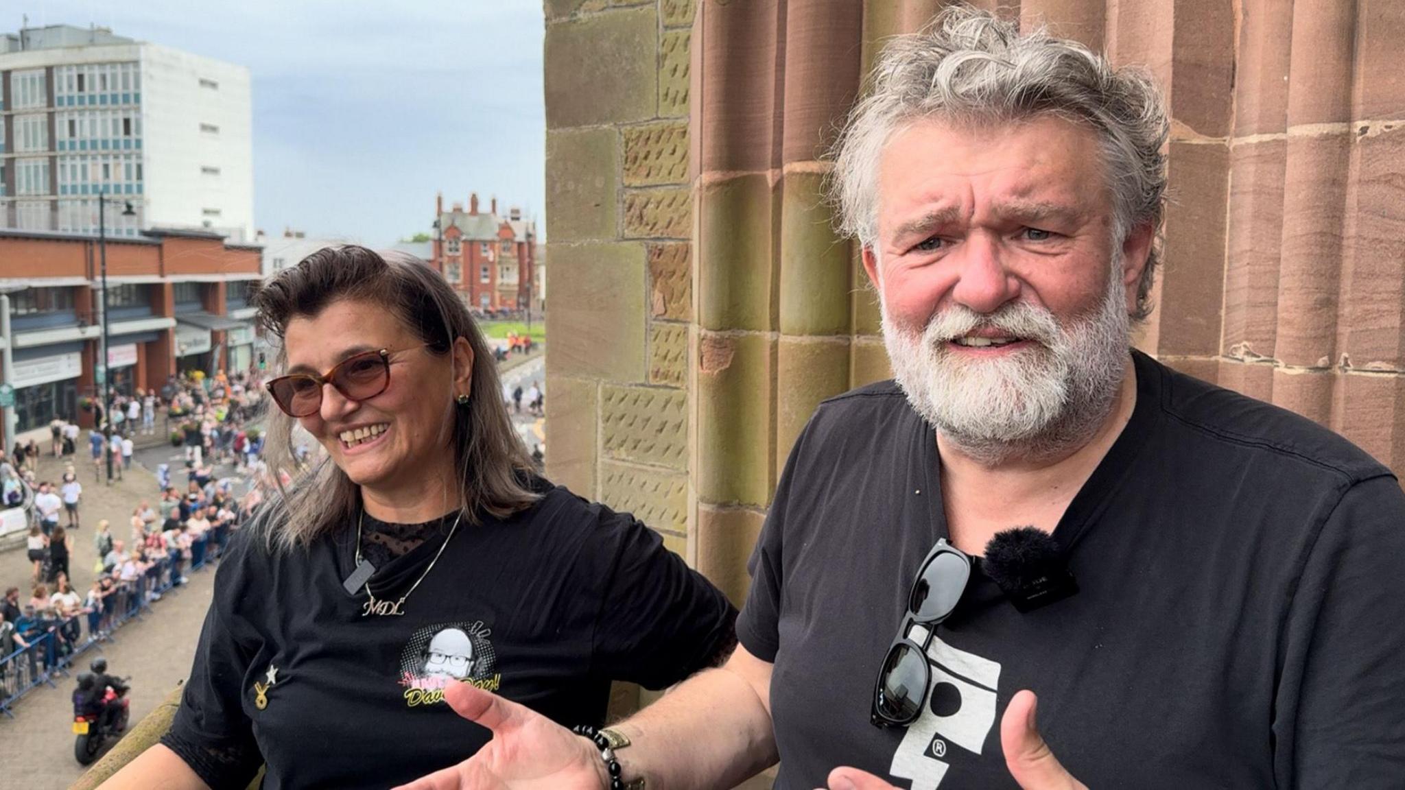 Lili Myers and Si King, who are wearing black t-shirts and sunglasses, stand on a balcony above Barrow's high street, where there is a procession of motorcyclists. Si has a grey beard and hair.