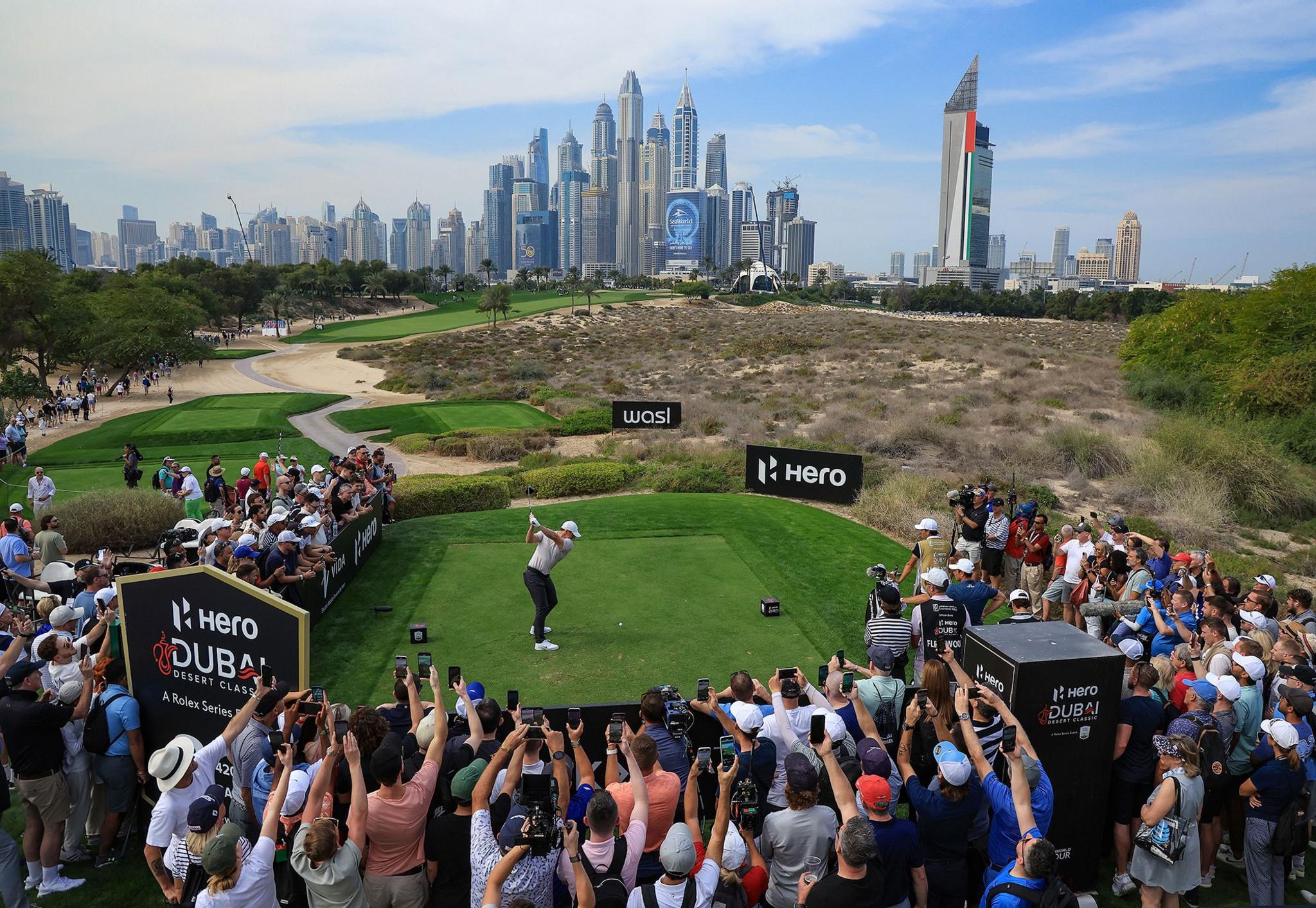 Rory McIlroy of Northern Ireland plays his tee shot on the eighth hole during the first round of the Hero Dubai Desert Classic at Emirates Golf Club. There are crowds surrounding the tee in the foreground and skyscrapers in the background