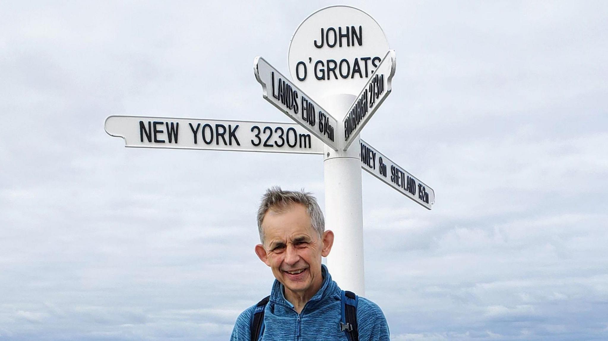 Prof Saunders stands below a white and black John O'Groats sign which has arrows pointing to New York, Land's End and Orkney and Shetland. He has grey hair and is wearing a blue jumper. 