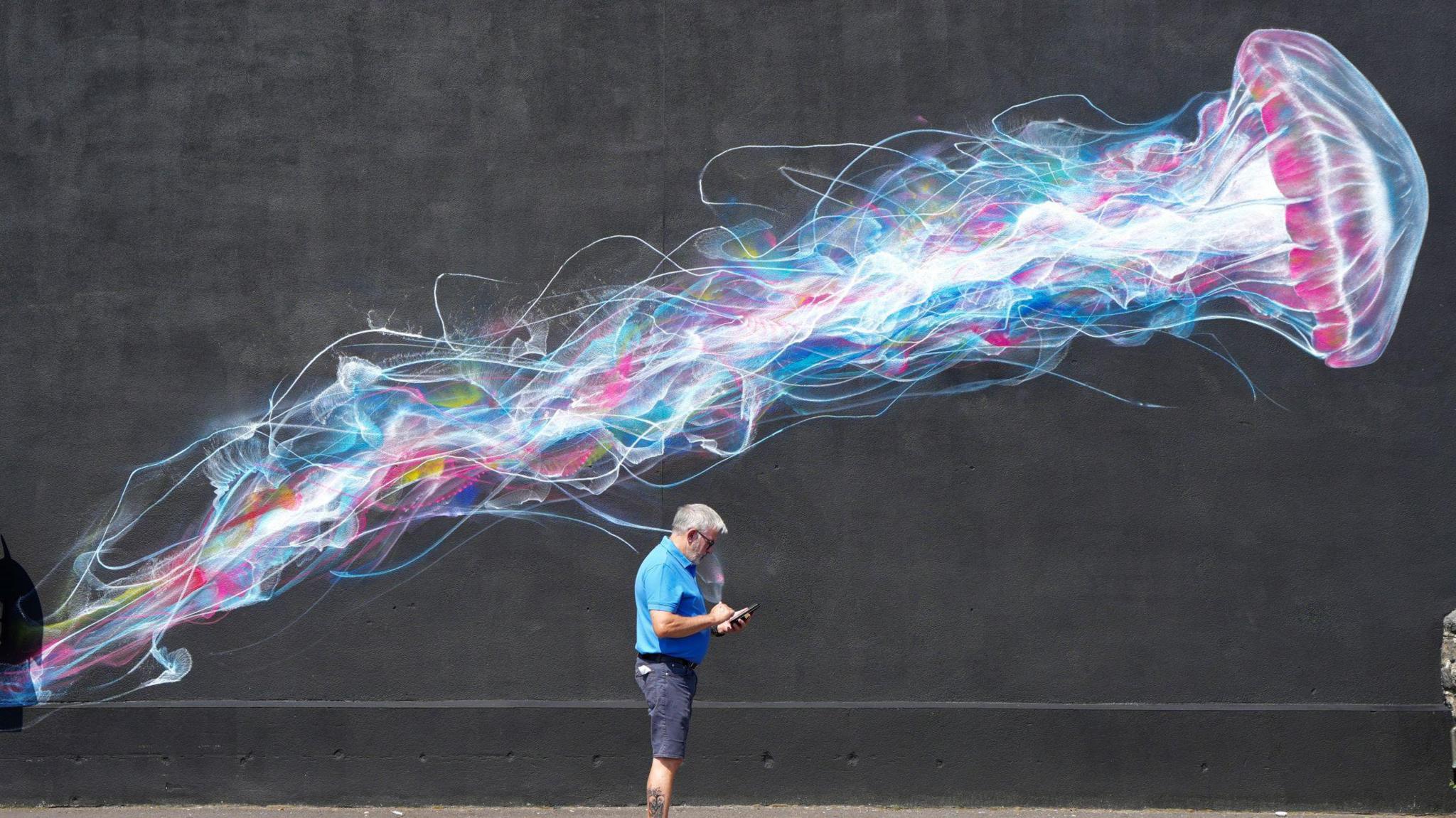 A man wearing blue denim shorts and a light blue T-shirt stands under a large street art mural in Weston-super-Mare. The mural is a multicoloured jellyfish stretching from left to right on a black background. The man is side on to the camera and looking down at his phone.