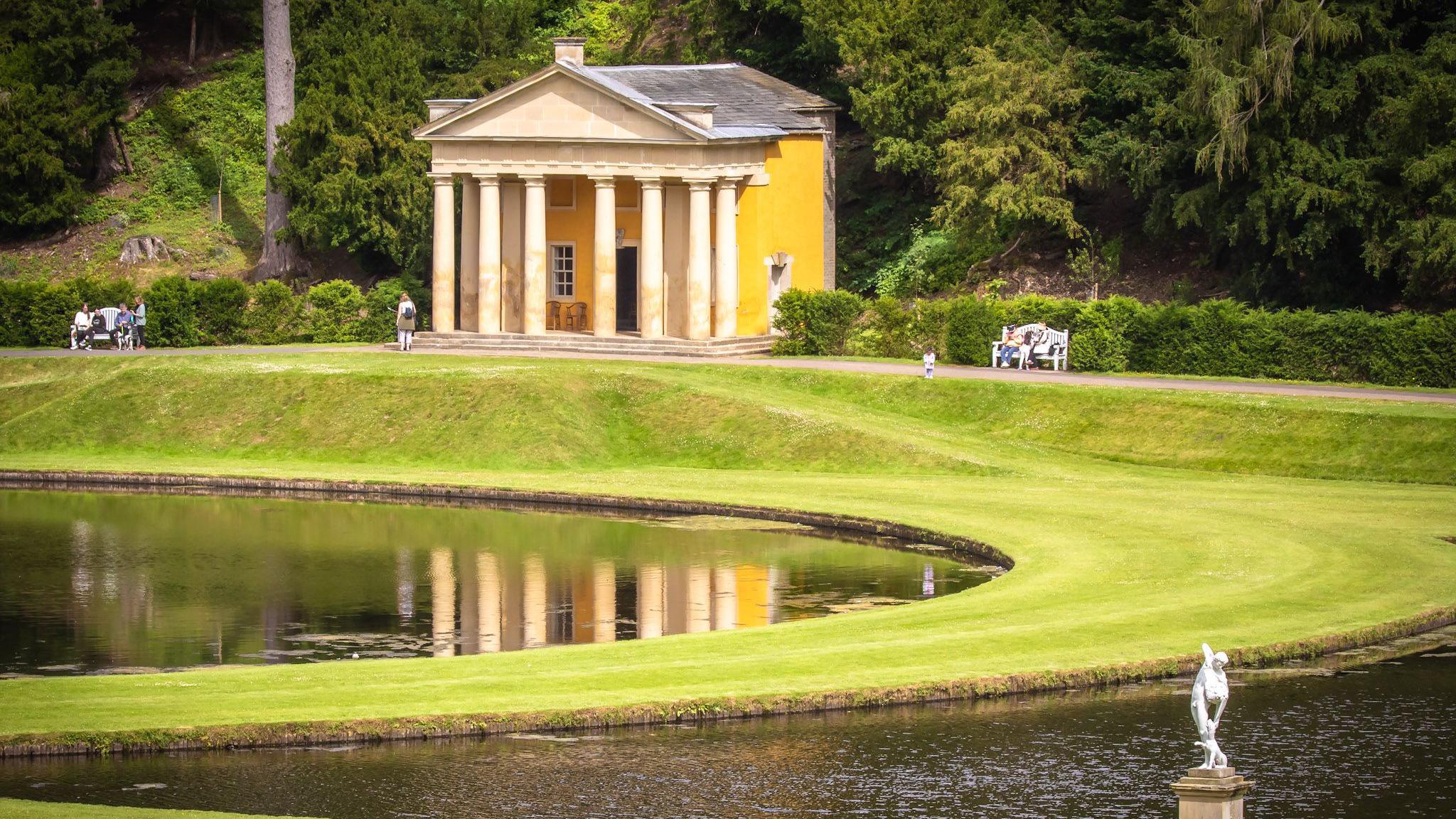 A view across a lake and manicured lawns, with stone statues and a Greek temple-style building