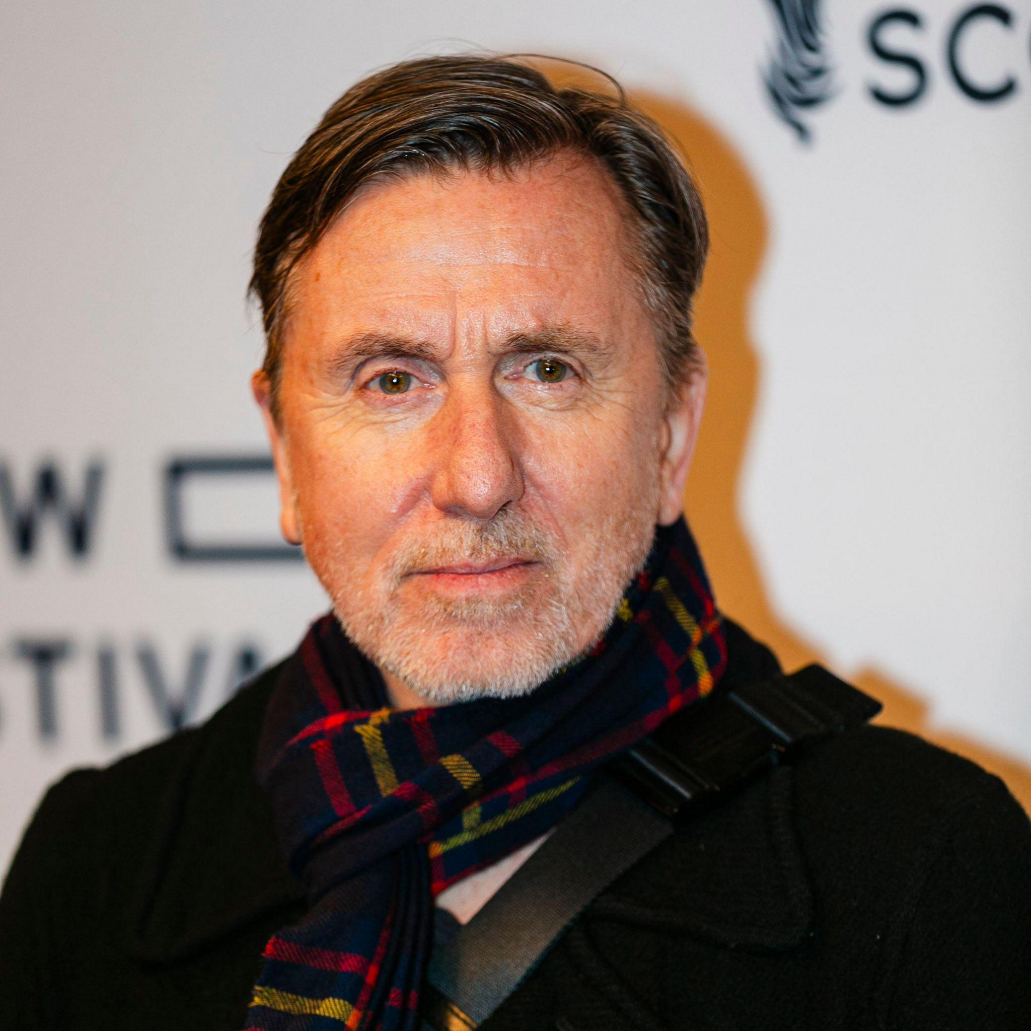 Tim Roth has brown hair and a short greying beard. He is wearing a dark jacket and a tartan scarf