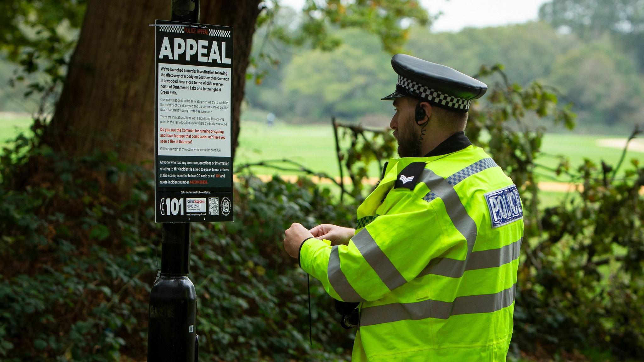 A police officer wearing a yellow hi-viz jacket and cap is attaching a poster to a lamppost. At the top of the poster it says Appeal. The rest of the writing is too small to see in the photo.