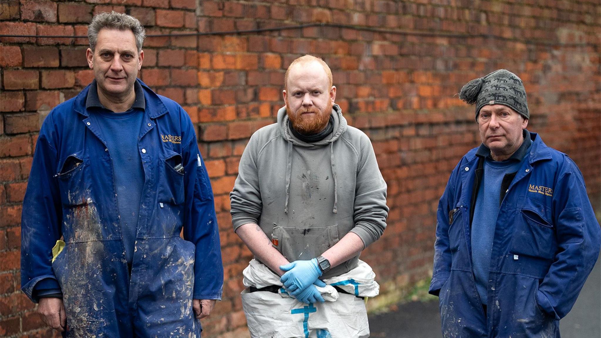 Three garage workers in their work clothes standing in front of a red-brick wall. Julian Medlock is the tallest, with greying hair and wearing blue overalls with paint and dirt from his work. Next to him is James Dixon, who has short red hair and a red beard, and is wearing a grey hoodie and his blue protective gloves. On the end is Colin Parry, who also wears blue overalls and has a grey woollen cap on.