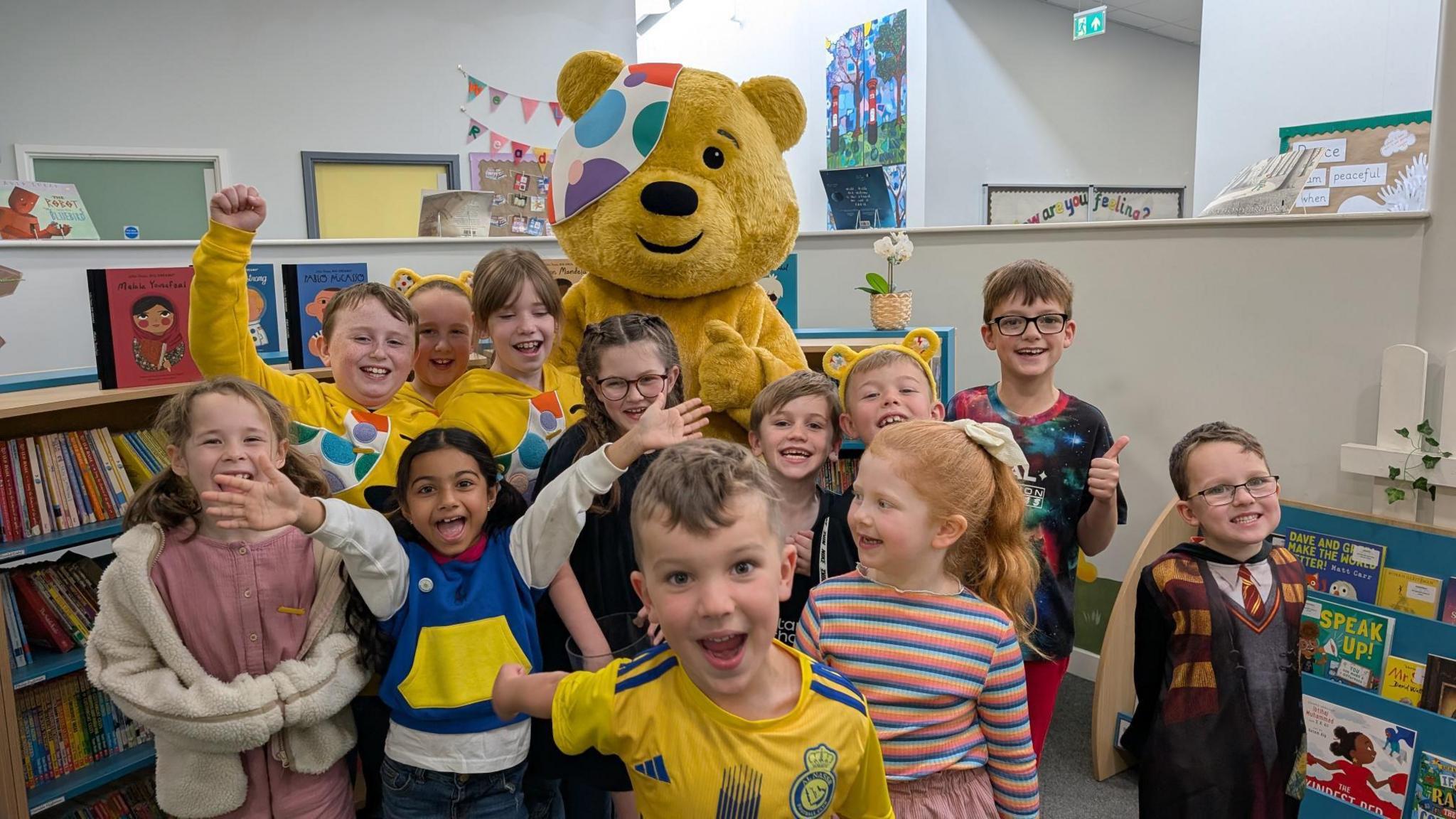 A class of children in a small school library all either smiling for the camera or cheering with Pudsey at the back with his thumbs up.