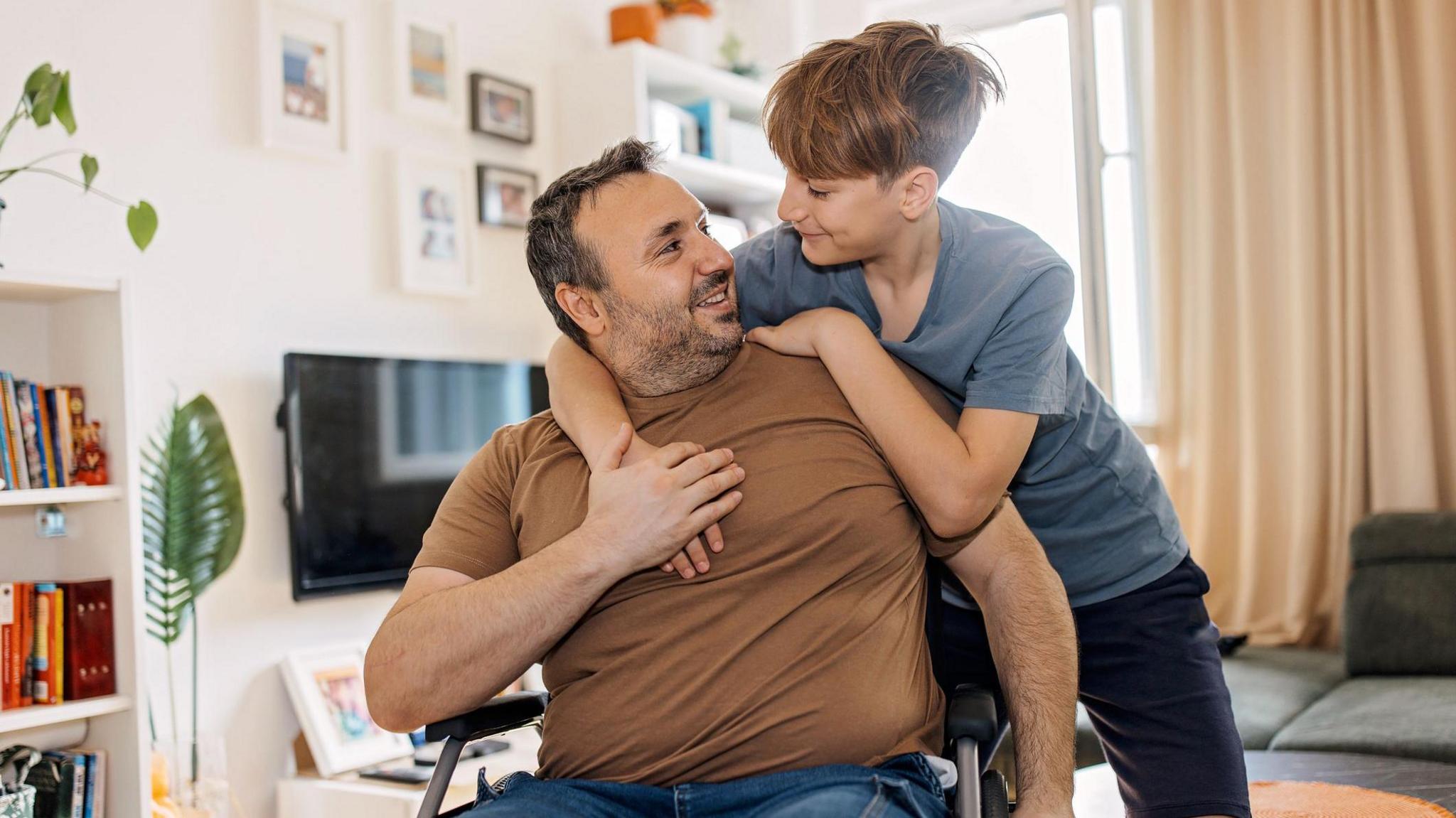 A boy gives his father, who is a wheelchair user, a hug in their home