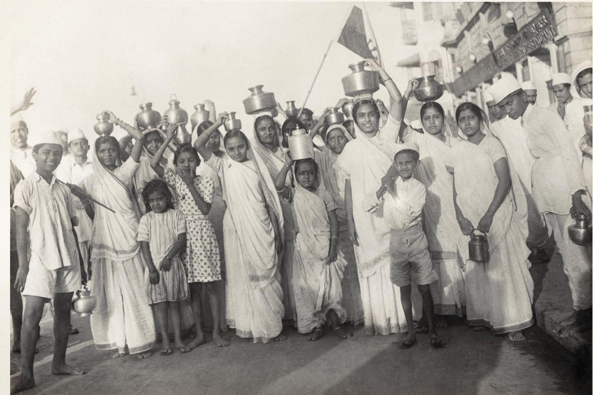 The monochrome photo shows a group of children, many of whom are carrying steel pots filled with sewater on their heads. The boys are dressed in loose shirts, shorts and caps, while the girls are wearing saris.