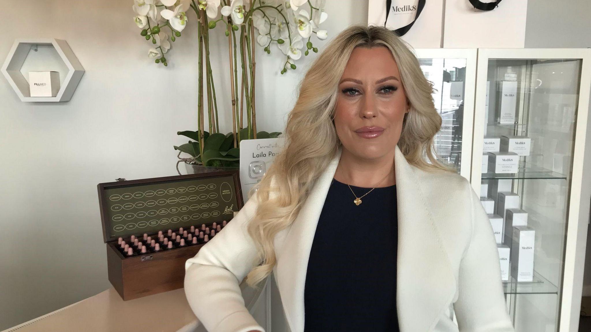 Owner of Laila Paris Aesthetic and Well-being Clinic in Horley, Melissa Farry, pictured in her store. A display unit with products can be seen in the background alongside a flower design and a reception table that Melissa is leaning on. Melissa has curled blonde hair and is wearing a white blazer jacket and black top underneath.