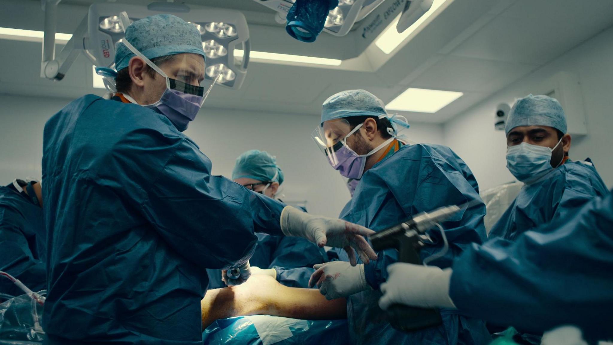 Doctors and nurses wearing blue scrubs and facemasks and gloves. They are carrying out surgery on a patient.