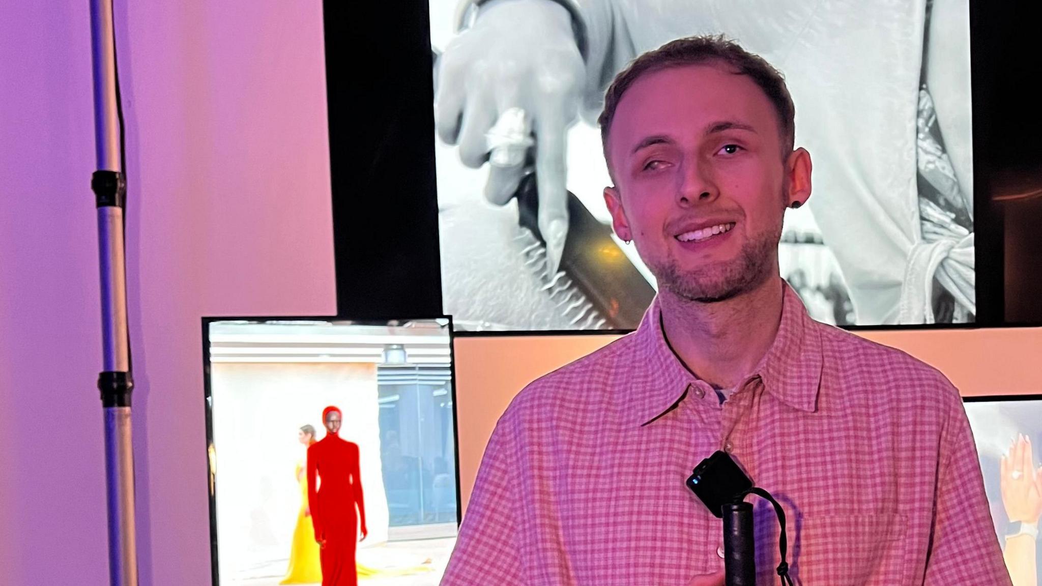 Aleks Tamasauskas at Chet Lo's London Fashion Week show. Mr Tamasauskas wears a pink and brown checked shirt and he holds a grey cane with a black top. He stands in front of three screens showing images from London Fashion Week