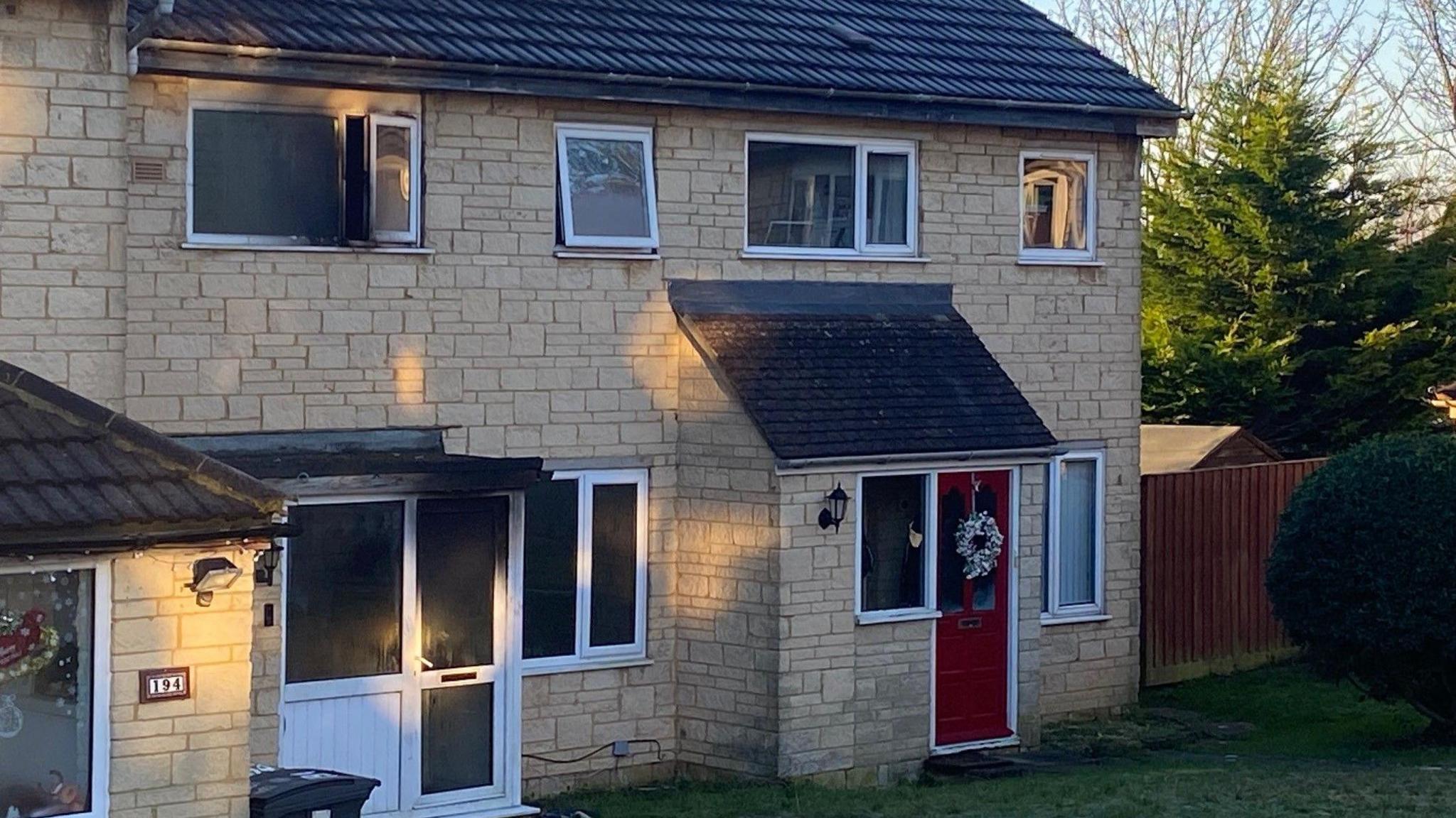 Two houses, a mid-terrace and a semi-detached, built out of light coloured stone, on a sunny winter's day. The mid-terrace house has soot all over one of its upstairs windows, and all over its white front door and porch.