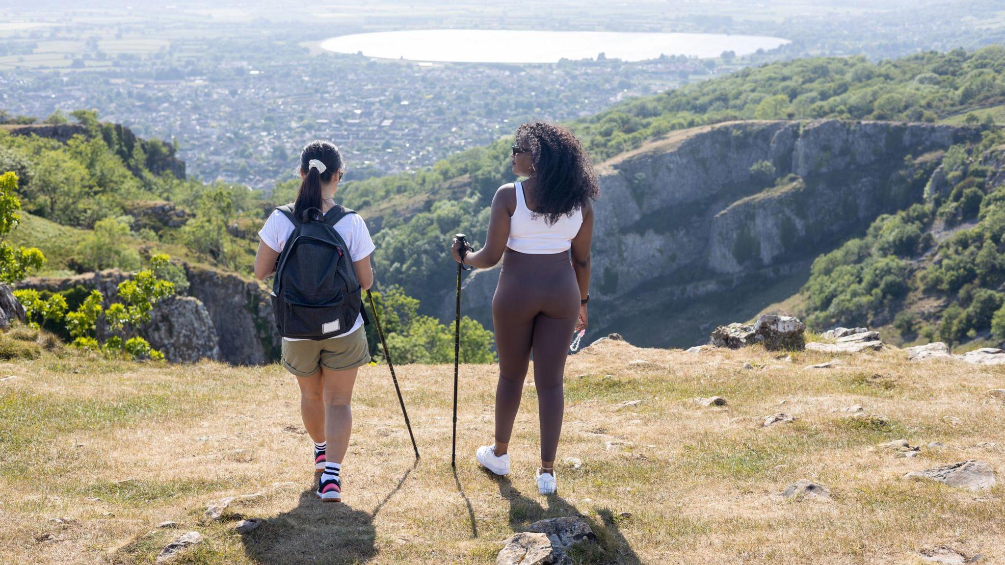 Two women dressed in walking gear and holding walking poles stand at the top of a hill overlooking a village and a reservoir on a summer's day.
