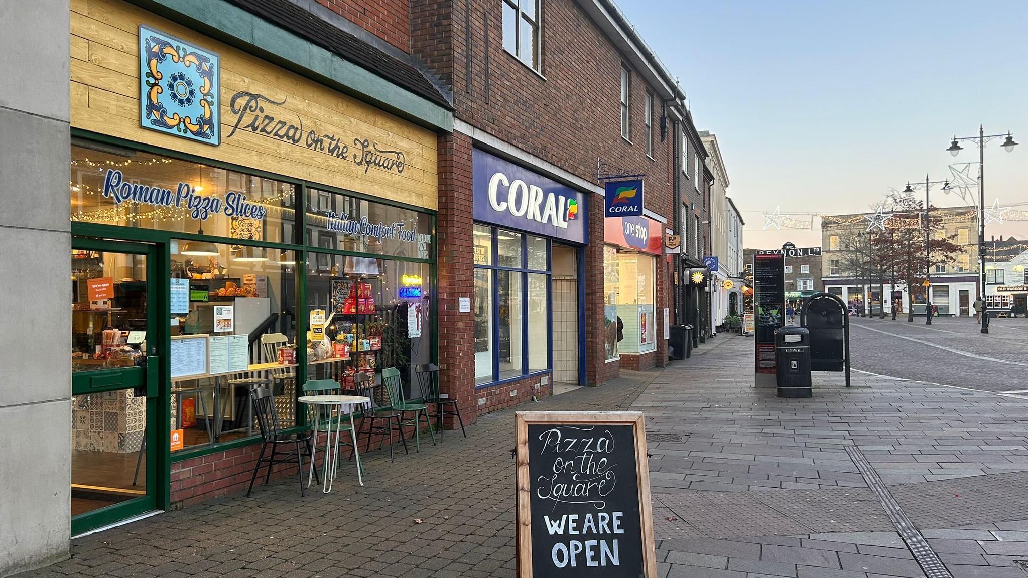 An outside image of the pizza on the square restaurant on a street next to other shops