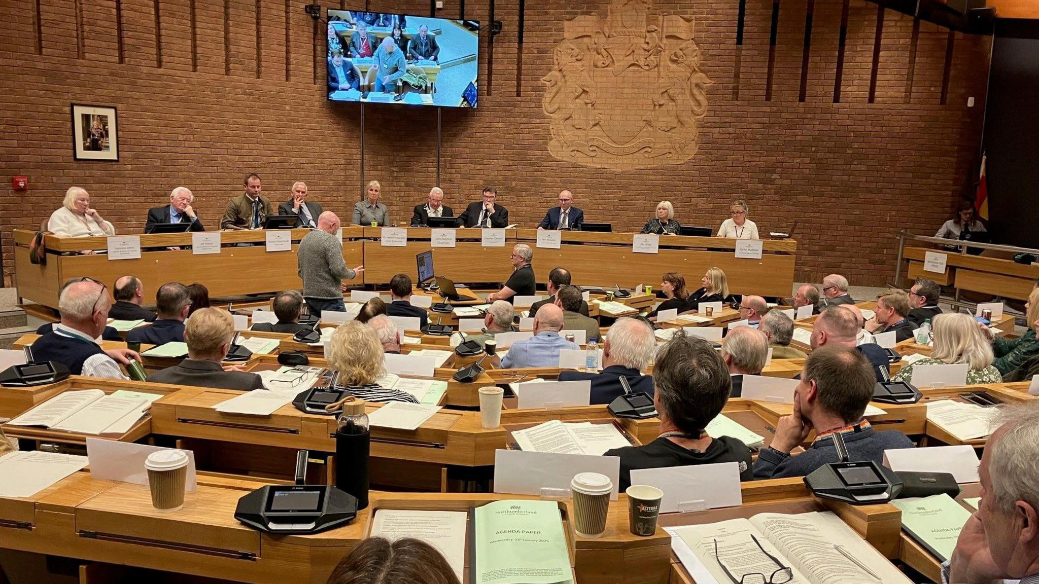 The inside of Northumberland County Council's council chamber showing someone talking and around 100 councillors sitting in a semi-circle listening. 