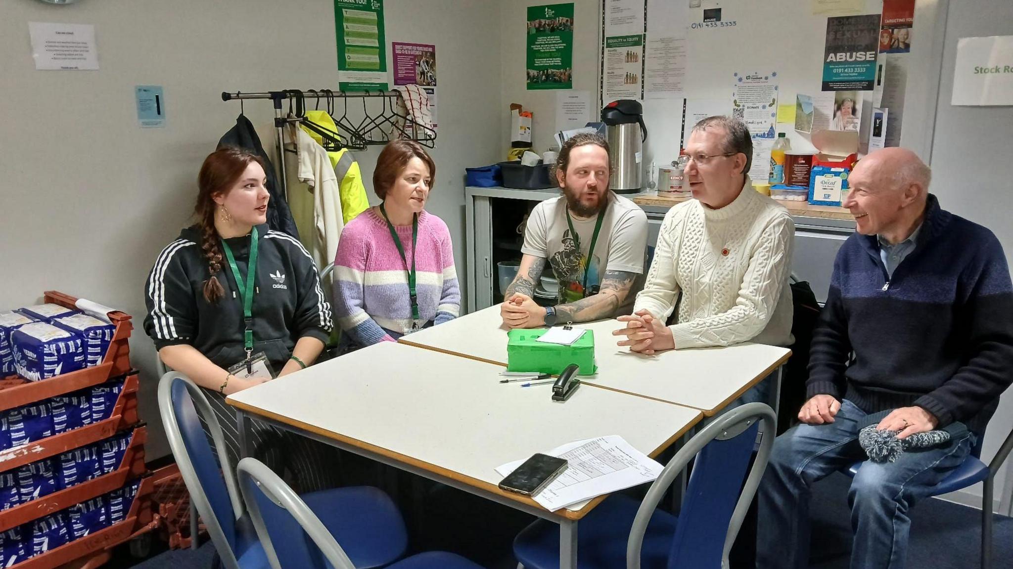 Foodbank volunteers and staff - two women and three men - in conversation as they sit around a table. Several loaves of bread are stacked next to a wall on the left-hand side of the room. 