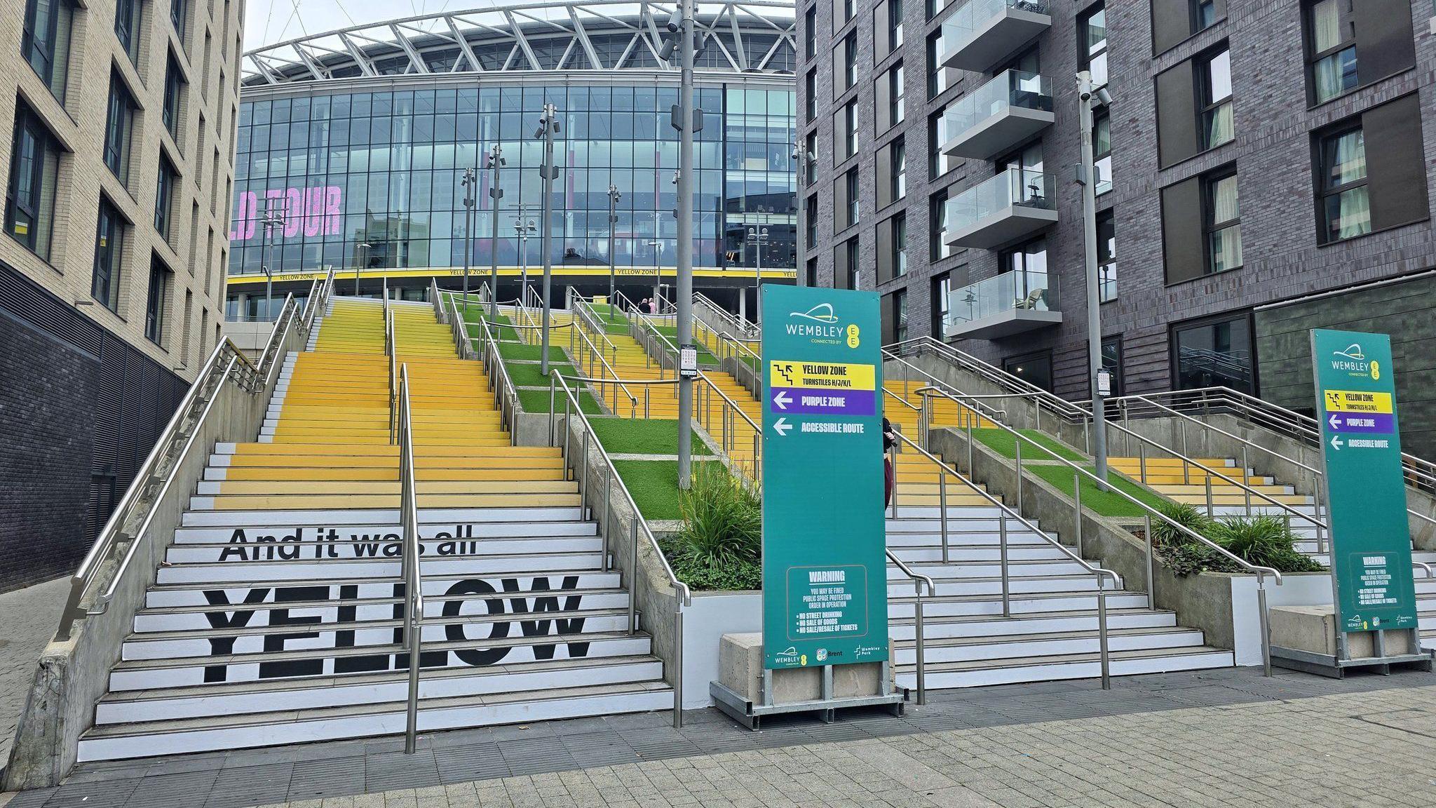 A set of steps leading the way up to Wembley Stadium, between two tower blocks. The steps are painted white, with different shades of yellow leading up the steps. In the bottom left, writing is painted in black reading 'And it was all yellow'.