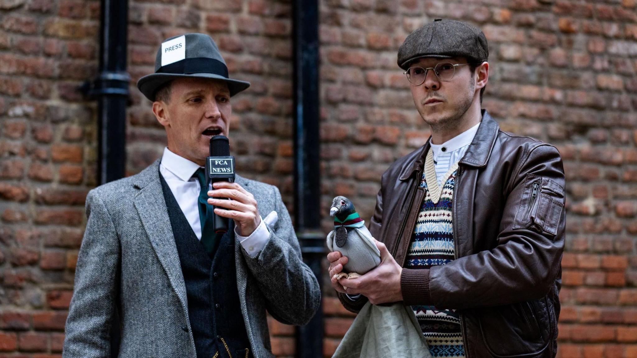 Two actors shown standing next to each other, with the man on the right clutching a fake pigeon. The man on the left is talking into a microphone with 'FCA News' written on it, and is wearing a press hat and a light grey suit. The man on the right with the pigeon is wearing 1940s-era dress, including a flat cap, and has a straight face. They are both stood in front of a brick wall.