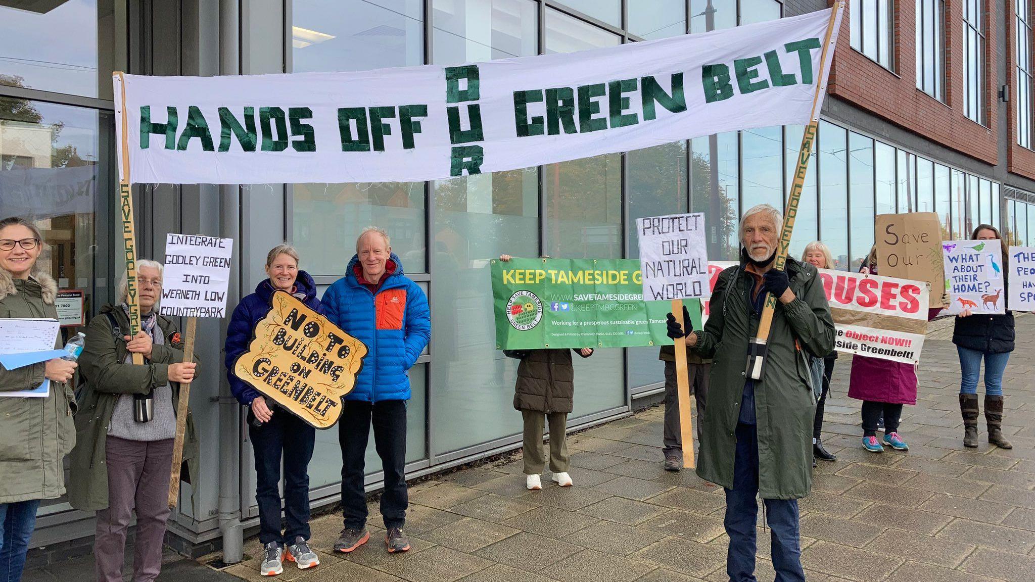 A group holding signs protesting the development. One is a large white banner with "hands off our green belt" written in green