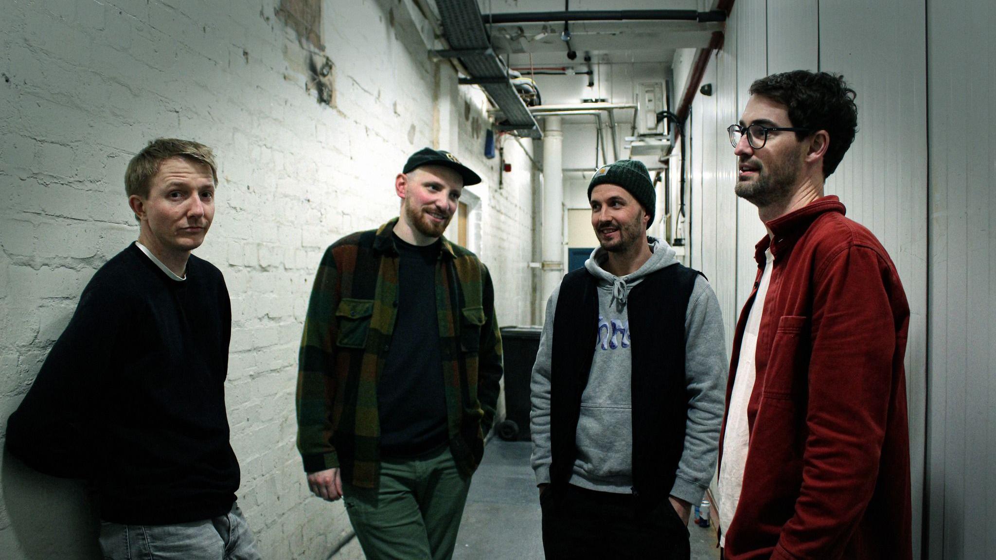 Four men stand in a corridor with white brick walls