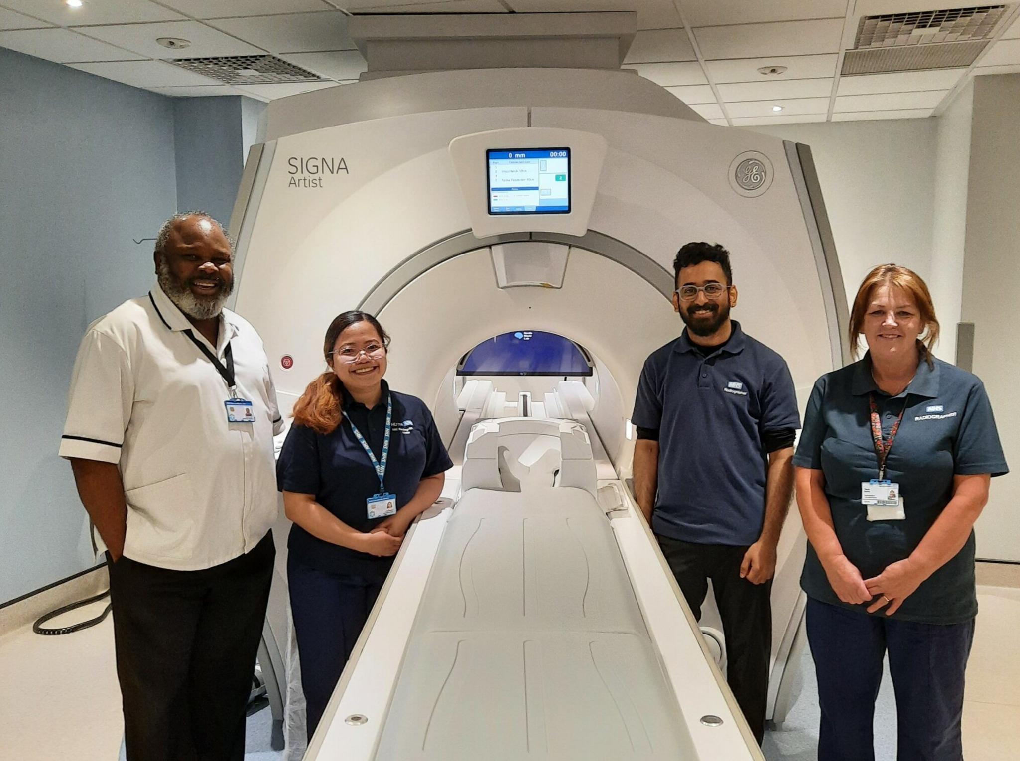 Four members of staff stand around an MRI imaging machine. The trolley where a patient would lie, is outside the actual machine, which has a small circular entrance.