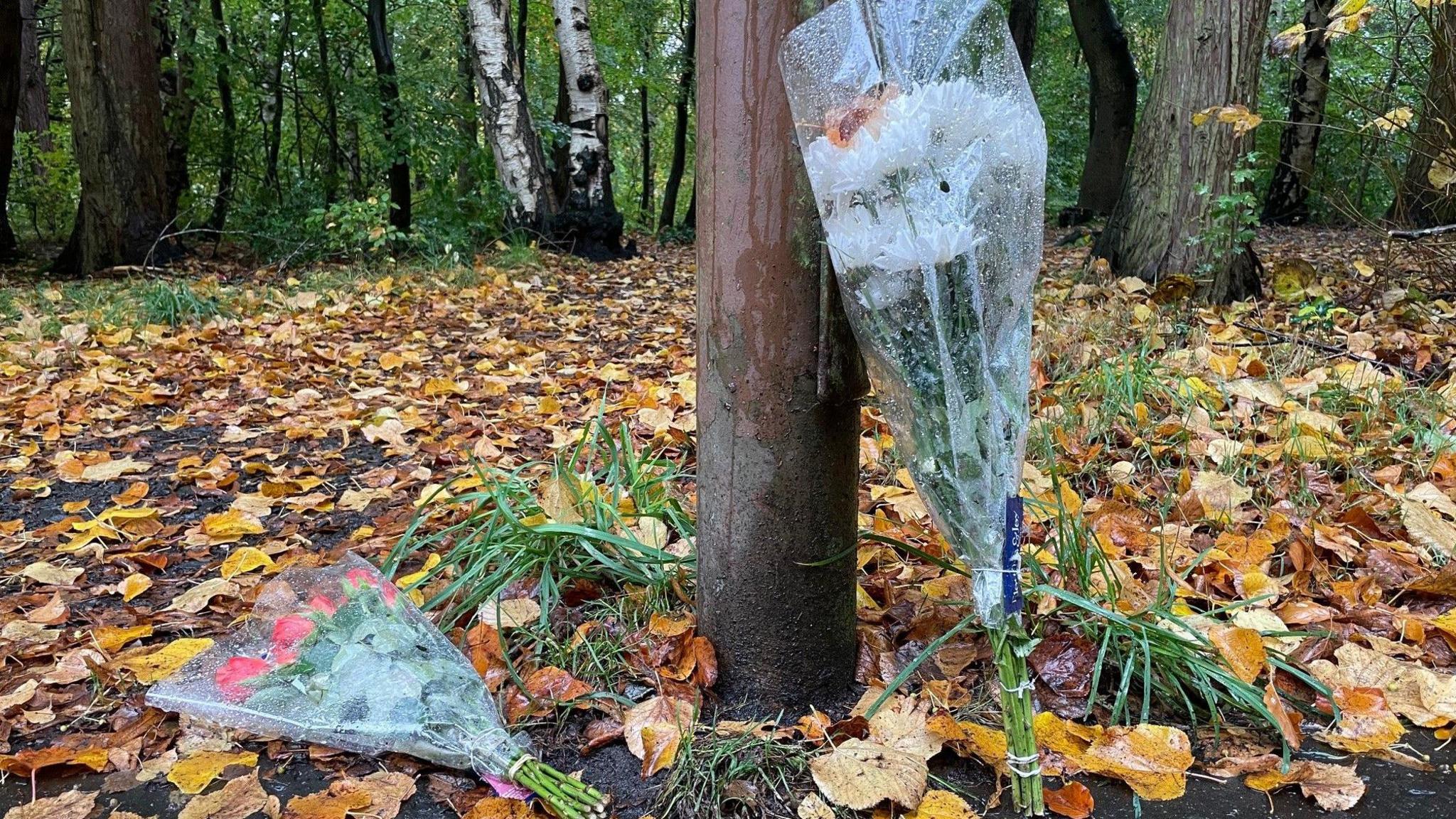 Floral tributes left leaning against a tree in autumnal woodland with orange, yellow and brown leaves on the floor in West Lothian near to the location of where Mr Nicol's body was found.