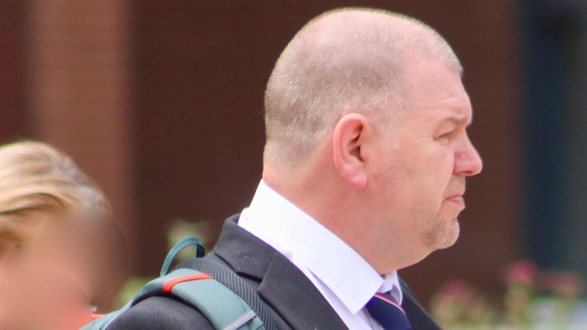 A side on view of Ian Ashworth, who has a shaven head and light stubble, walking away from court wearing a dark suit with a white shirt and blue and red striped tie.