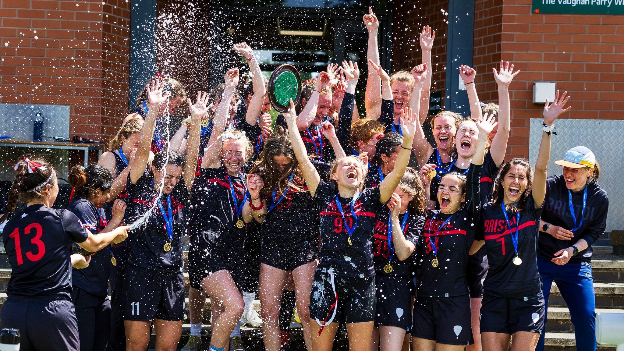 A group of young woman in dark blue and red sports tops and shorts cheer and shout with their hands in the air as one of their team sprays them with champagne.