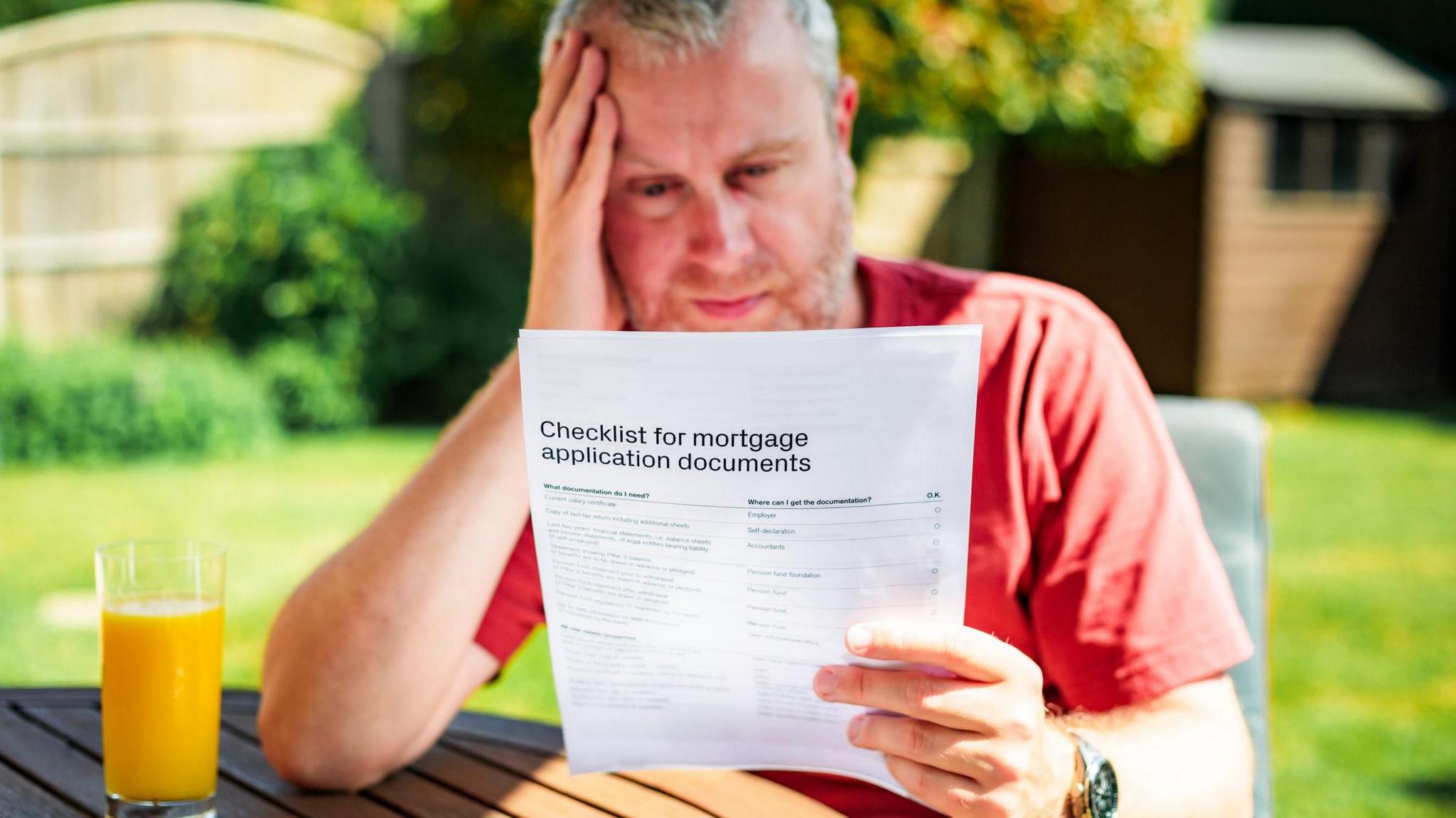 Man looks at mortgage application documents while sitting at a garden table with a glass of orange juice in front of him. There is a lawn and shed in the background. 