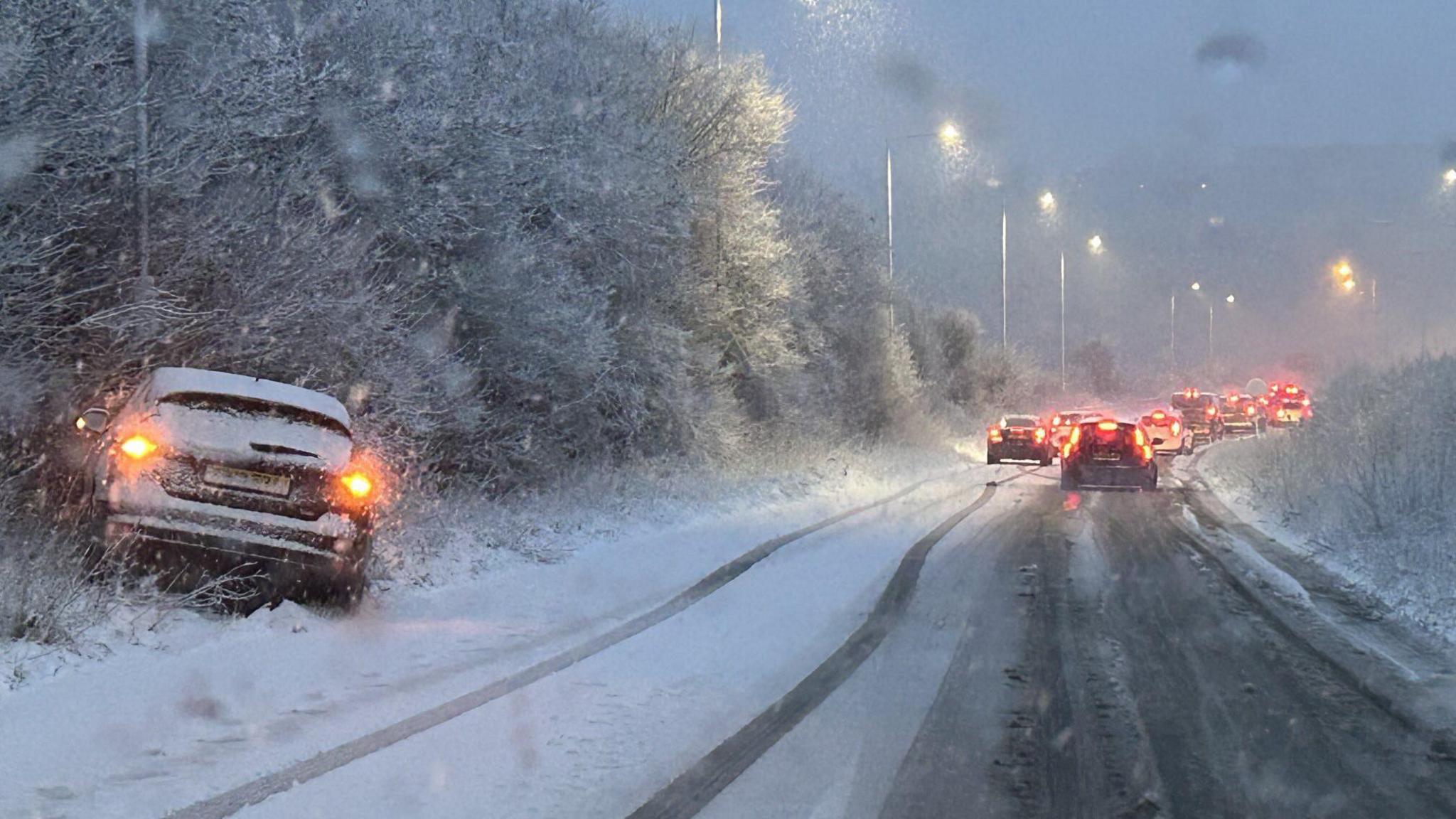 One car in snowy bushes on the side of the road and cars with brake lights on a slip road queue to join a carriageway. The road is covered in snow.