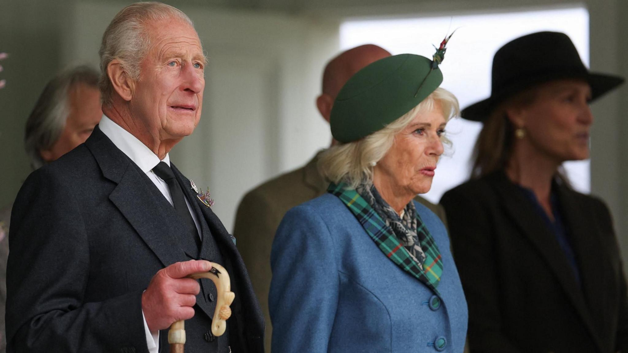 King Charles in a dark jacket and tie and white shirt is holding an ornate walking stick. Queen Camilla is wearing a blue coat with a tartan collar, green hat with a feather in it, and a scarf. They are pictured side-on