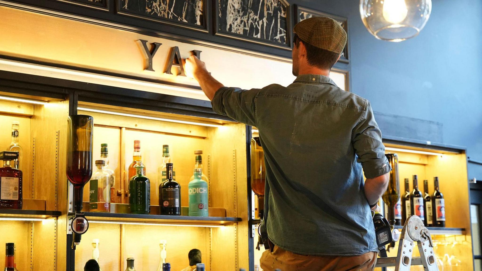 John Cooper paints a gold sign above a bar while standing on stepladders