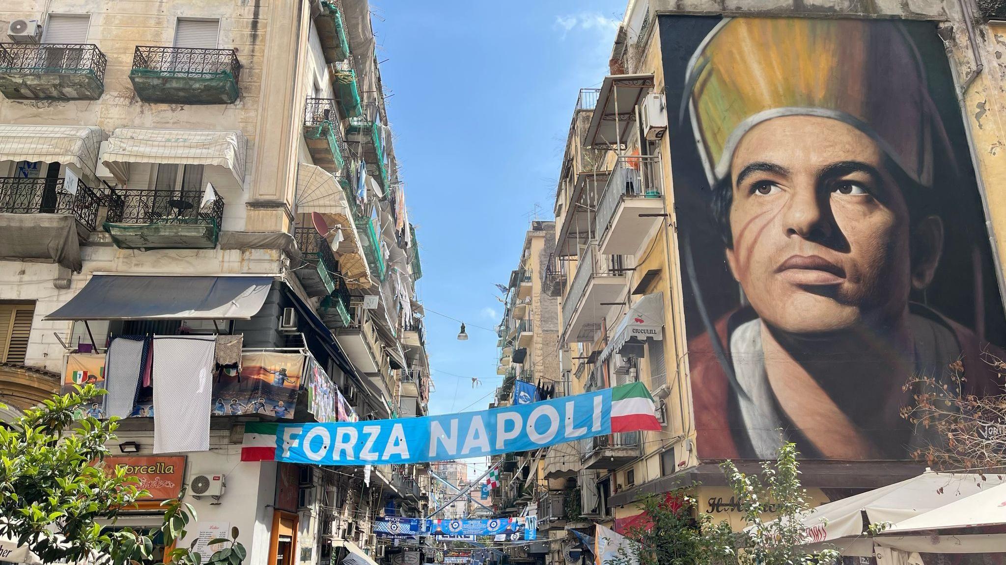 Huge wall mural in a Naples street. There are Napoli banners strung between flats on either side of the street