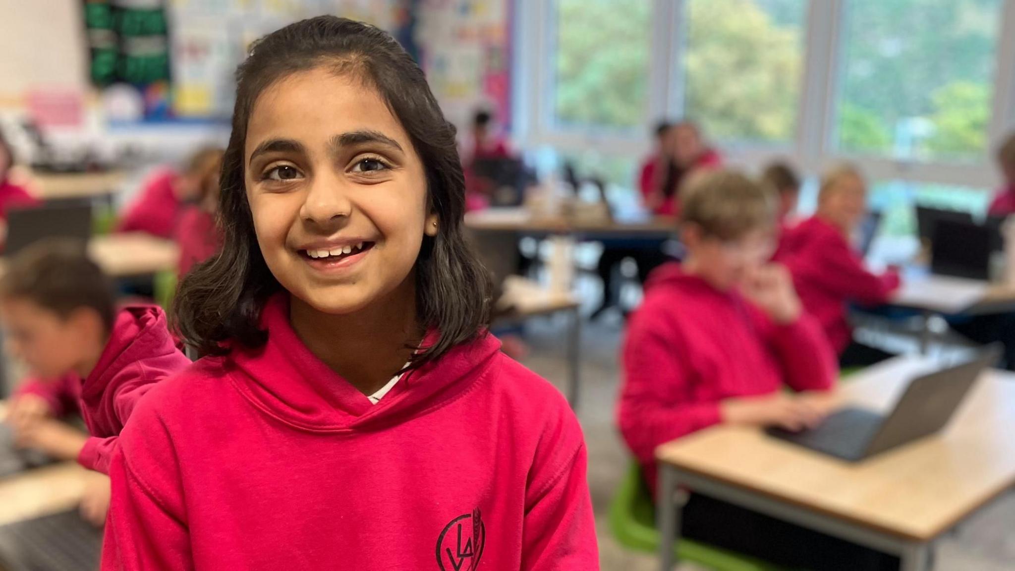 Aisha standing in a classroom with other pupils sitting at desks in the background. She is wearing a deep pink hoodie and smiling at the camera.